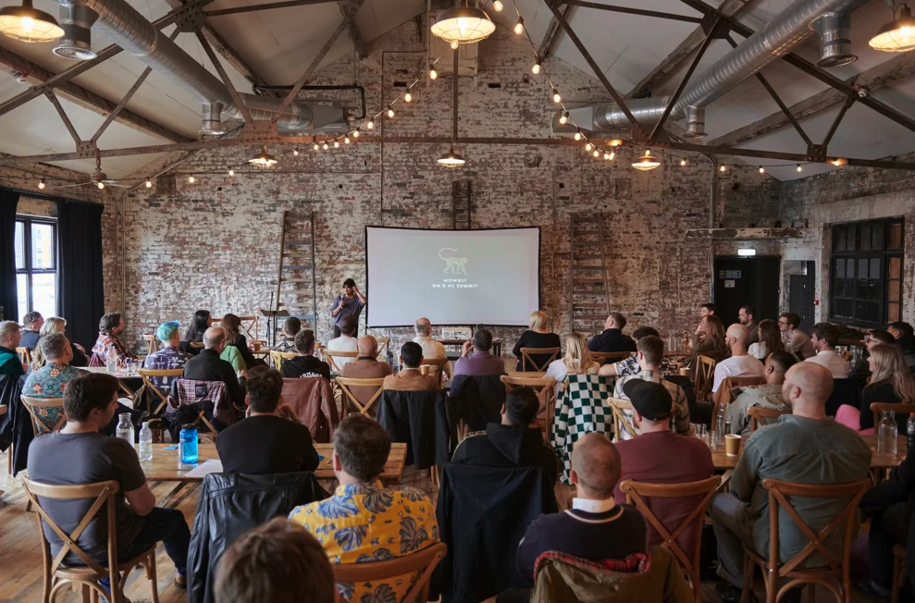 A large group of people is seated in a rustic space with a presentation screen at the front, while a speaker stands on stage engaging the audience.