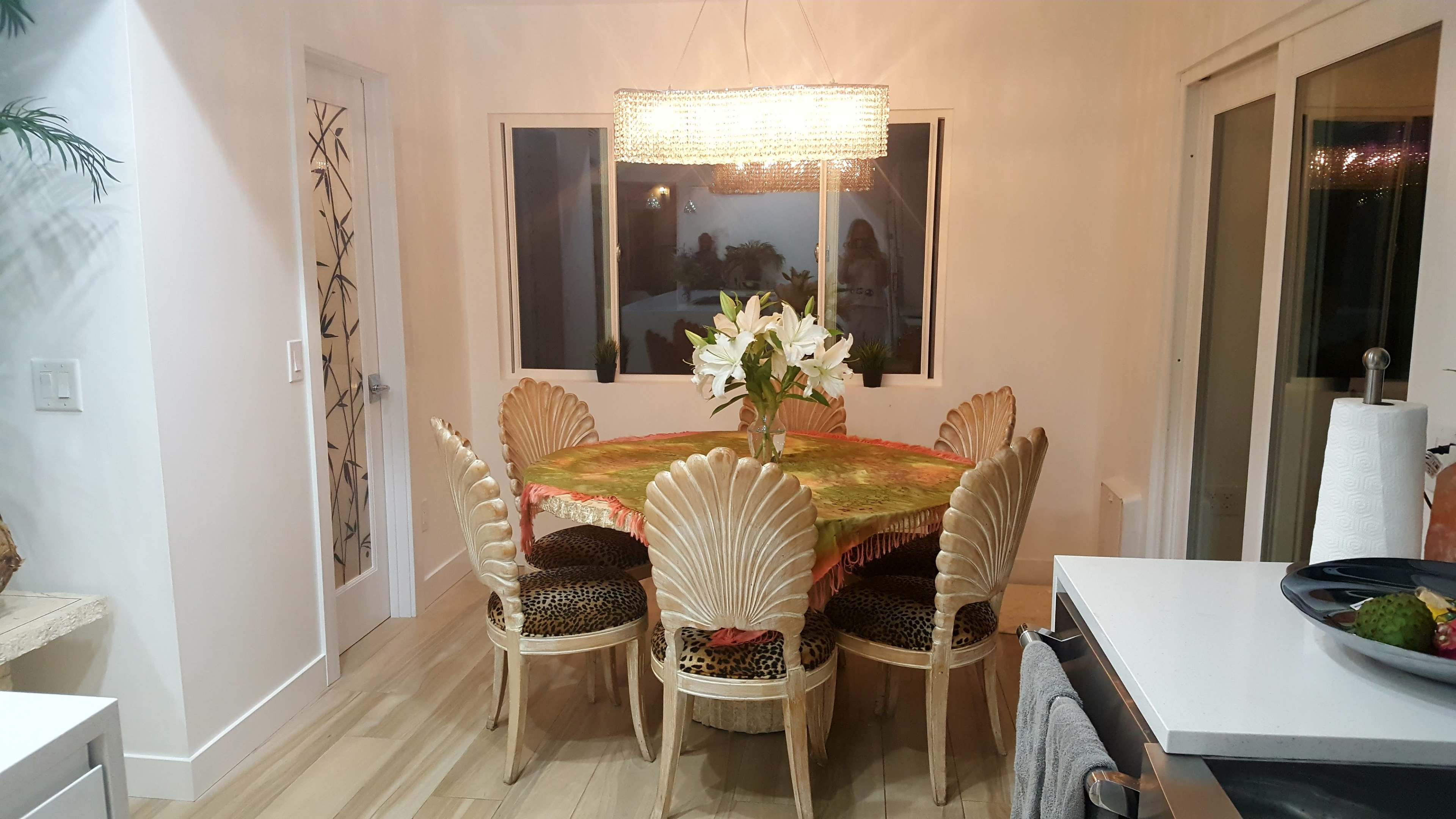 A round dining table with a colorful tablecloth is surrounded by ornate shell-patterned chairs in a well-lit room featuring large windows.