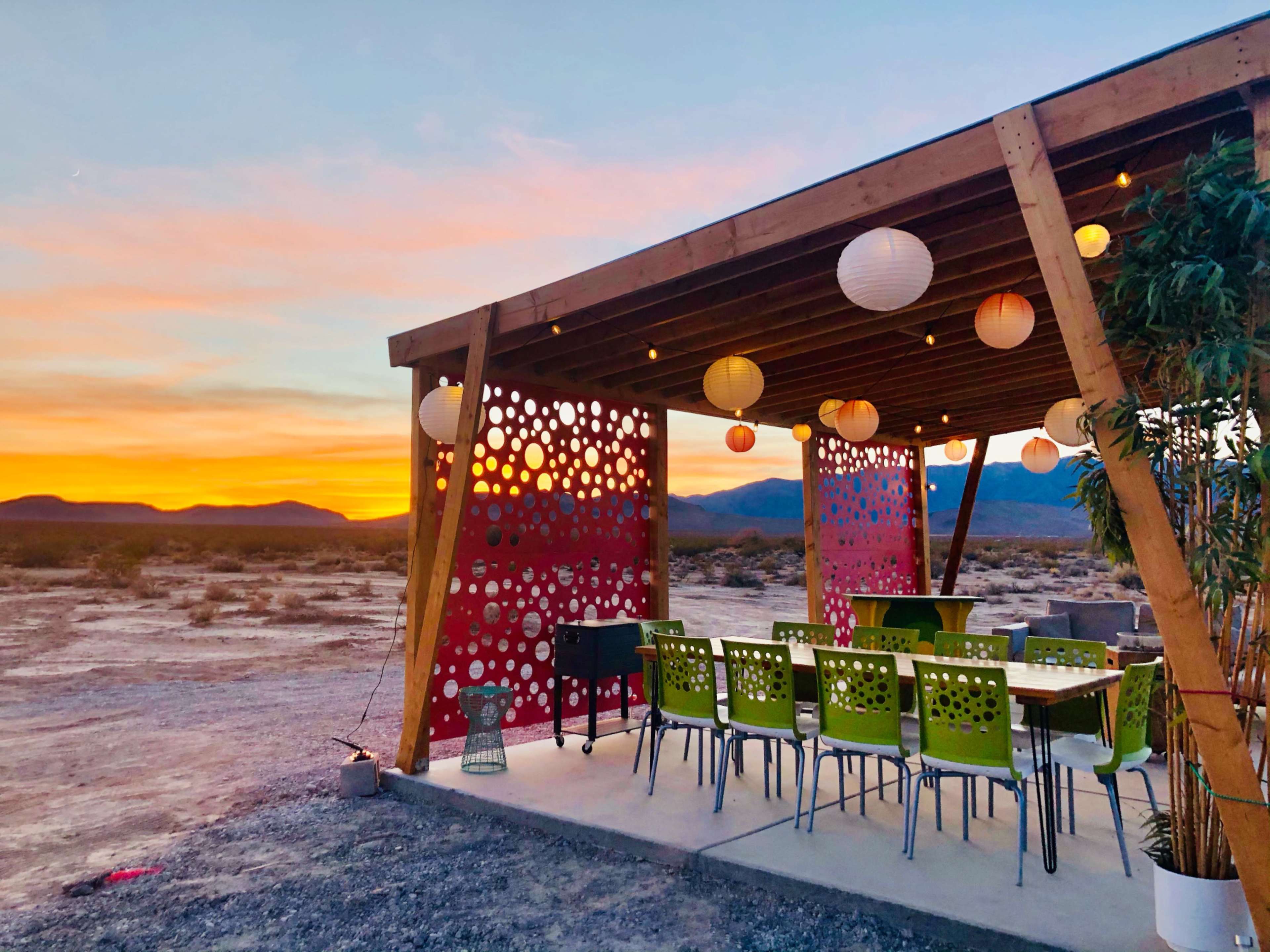A covered outdoor seating area with colorful chairs and hanging lanterns overlooks a desert landscape at sunset.