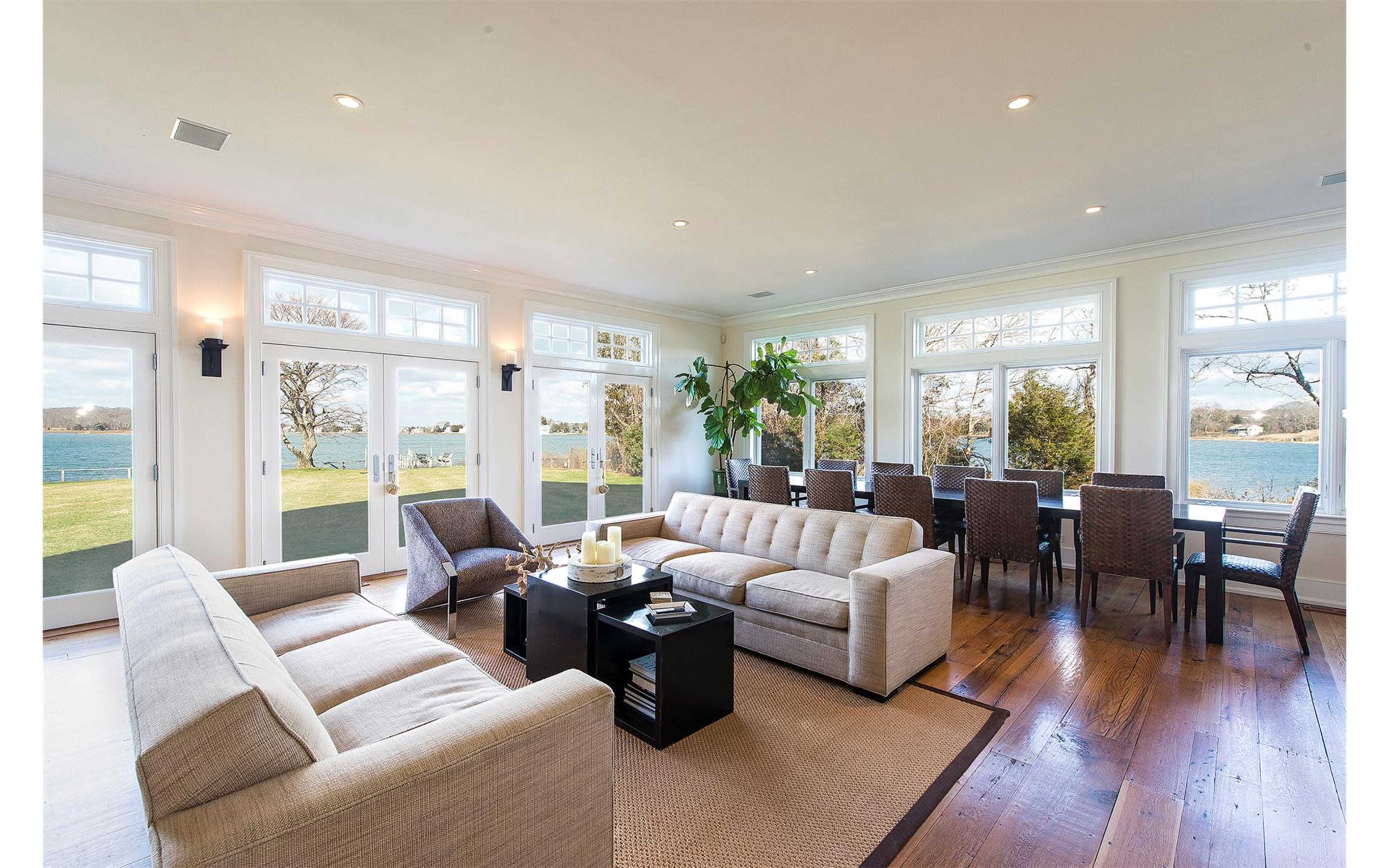 A spacious living room features beige sofas, a dark coffee table, and large windows overlooking a lake.