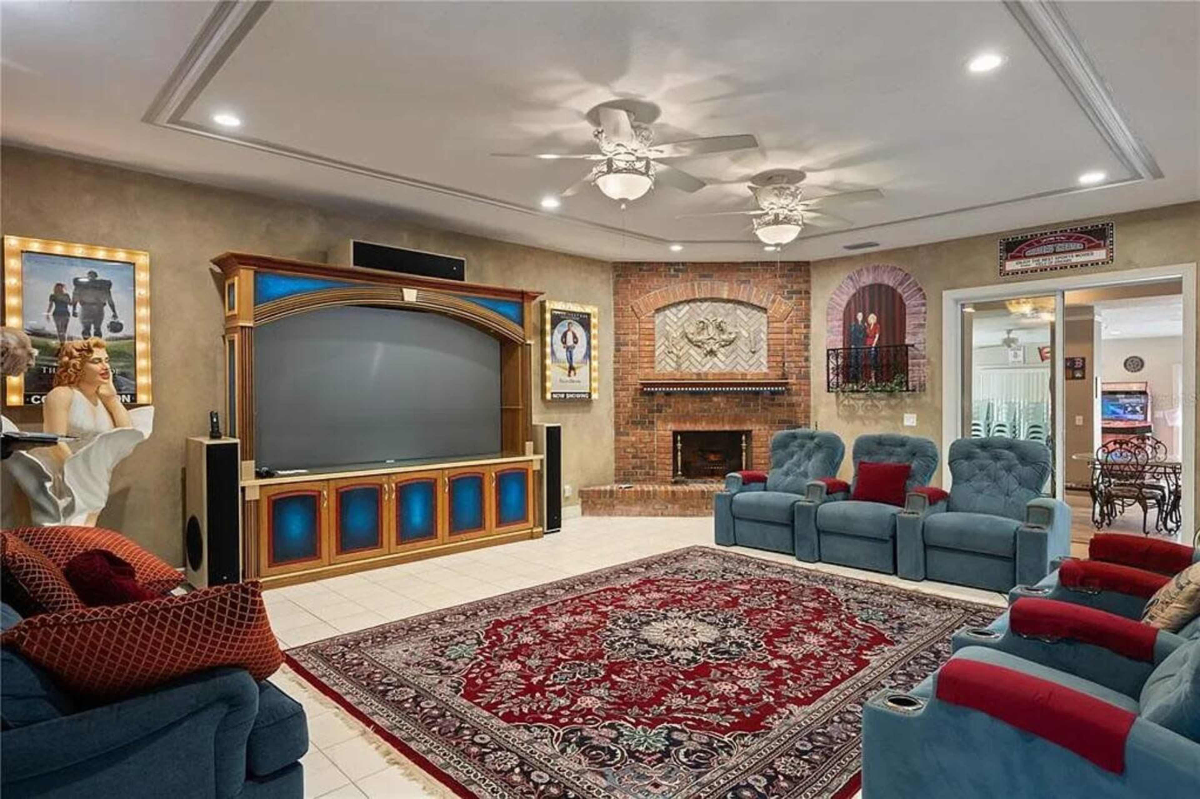 The image shows a spacious living room with a large entertainment center, blue recliners, a patterned rug, and a brick fireplace.