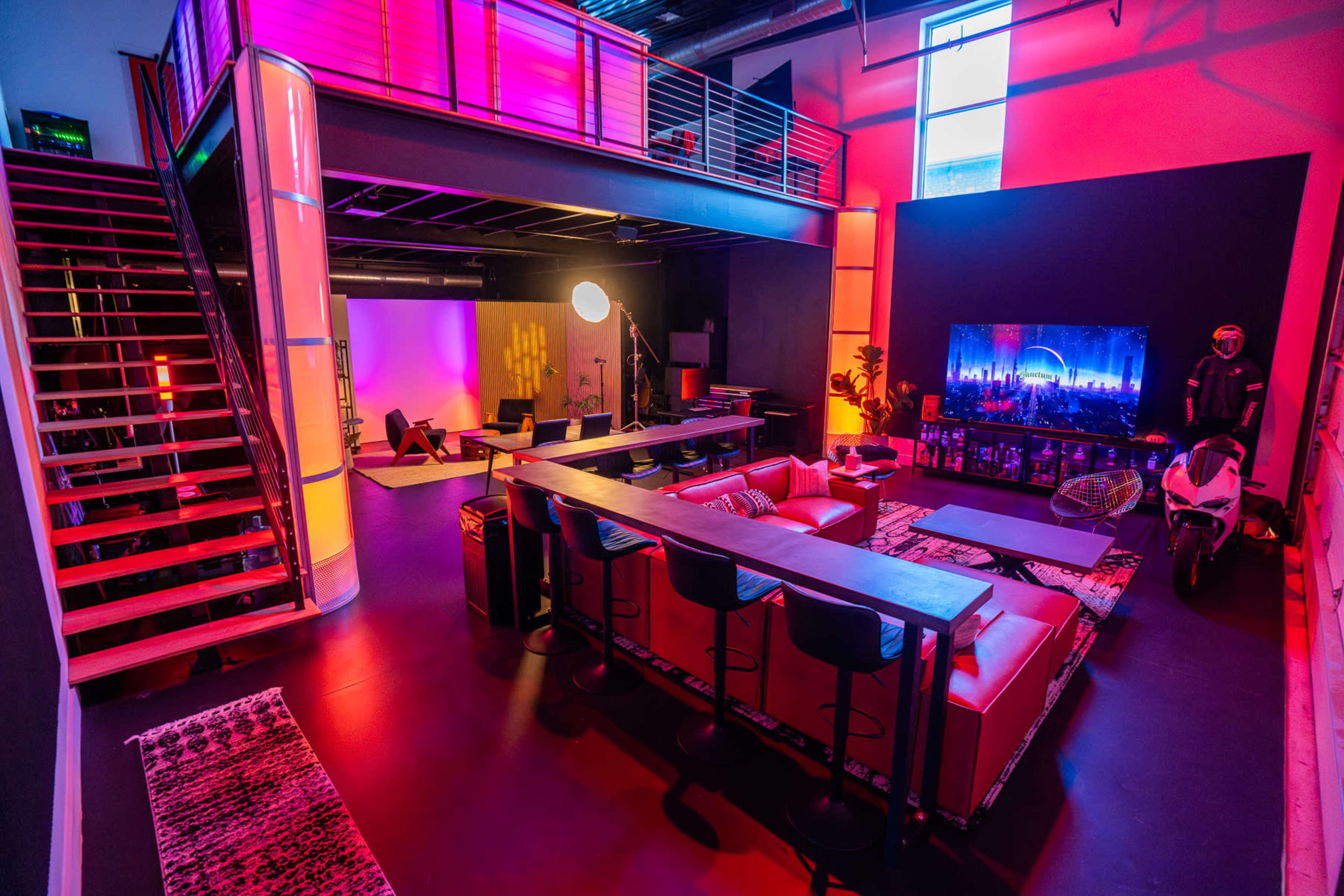 The image shows a modern, multi-level studio space with colorful lighting, a large seating area, a bar table, and a stairway leading to an upper level.