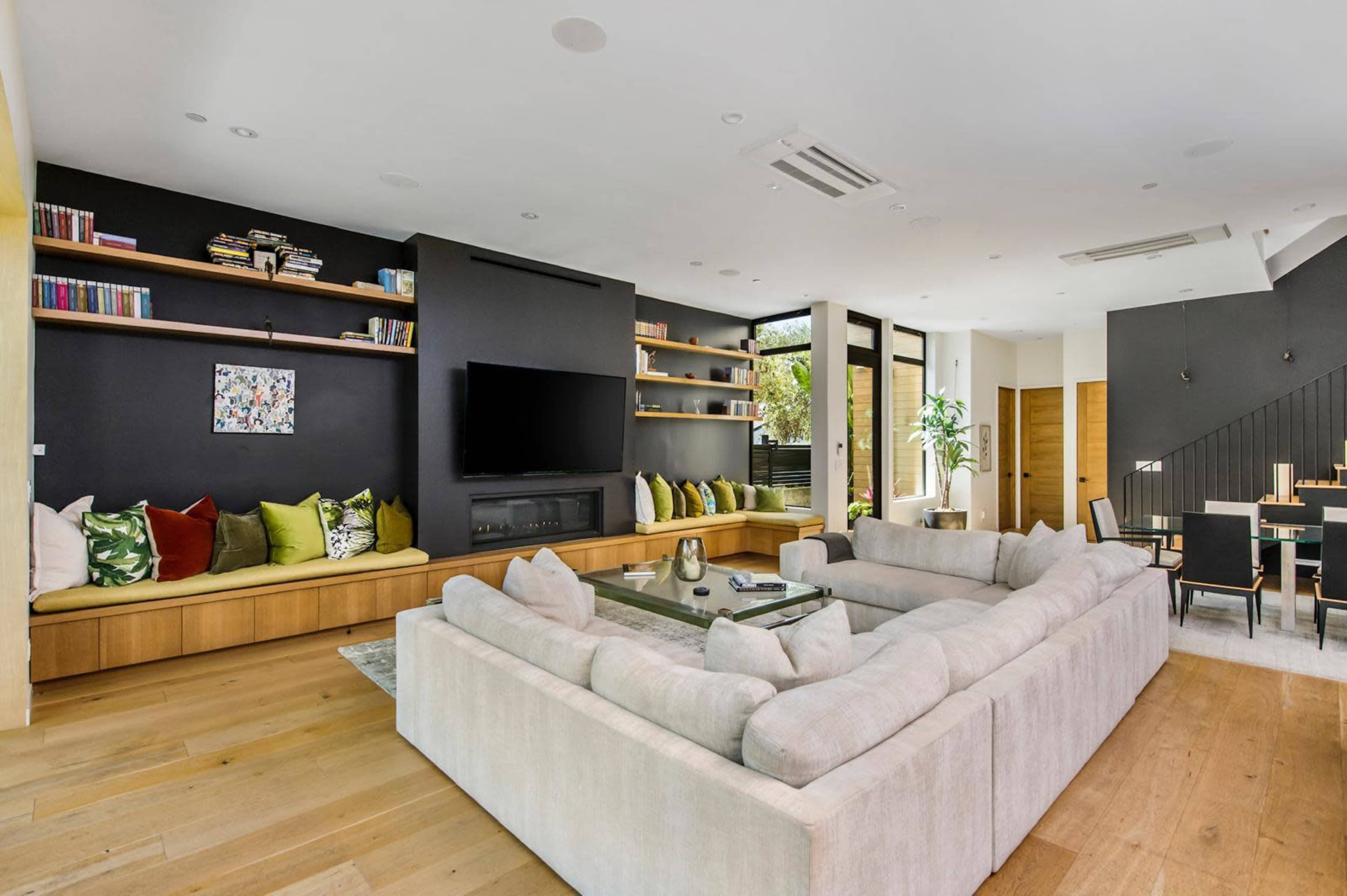 The image shows a modern living room featuring a large sectional sofa, a wall-mounted television, a shelf filled with books, and a dining area with a table and chairs.