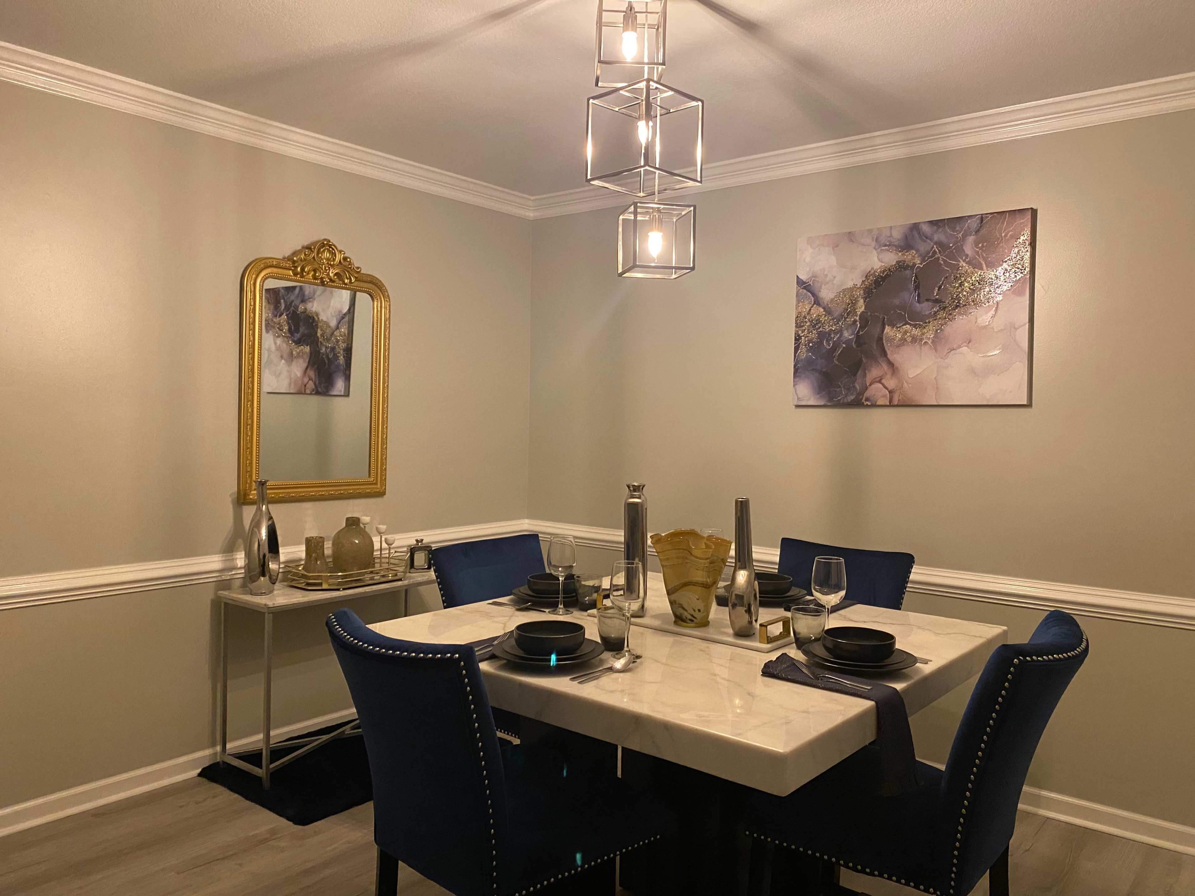 The image shows a dining area featuring a marble table set with plates and glasses, surrounded by blue upholstered chairs, with a gold-framed mirror and abstract artwork on the walls.