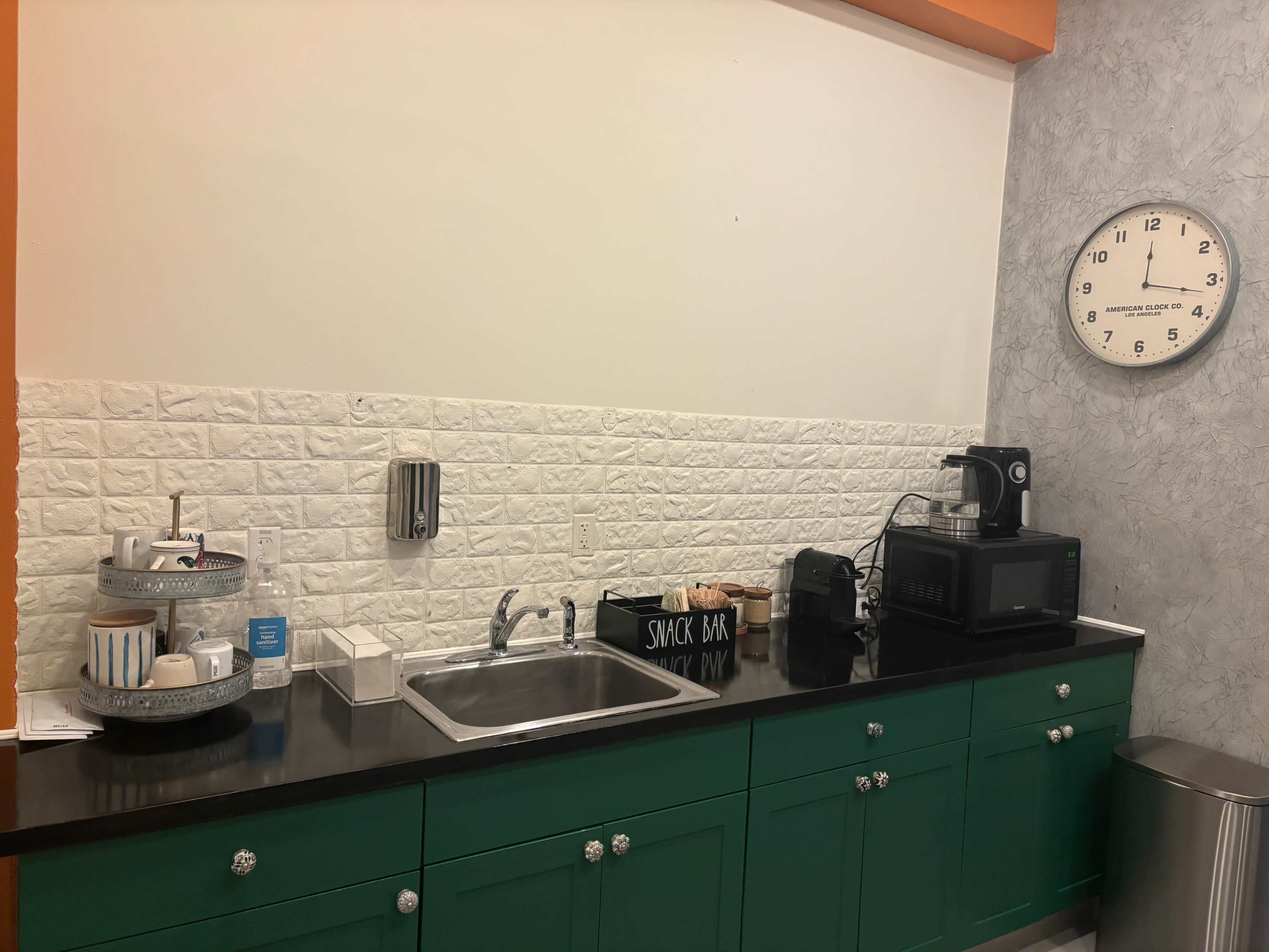 The image shows a kitchenette with a green cabinet, sink, microwave, coffee maker, and a clock on the wall.