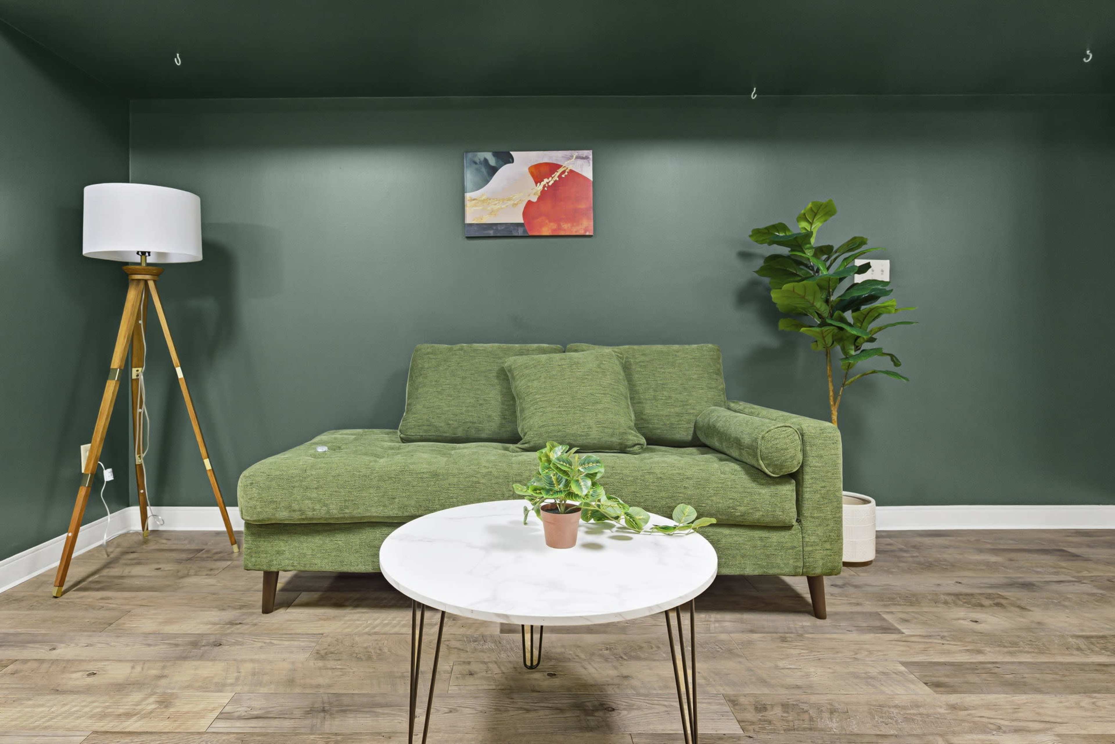 The image shows a small living area with a green sofa, a round white coffee table, a floor lamp, and a potted plant against a dark green wall.