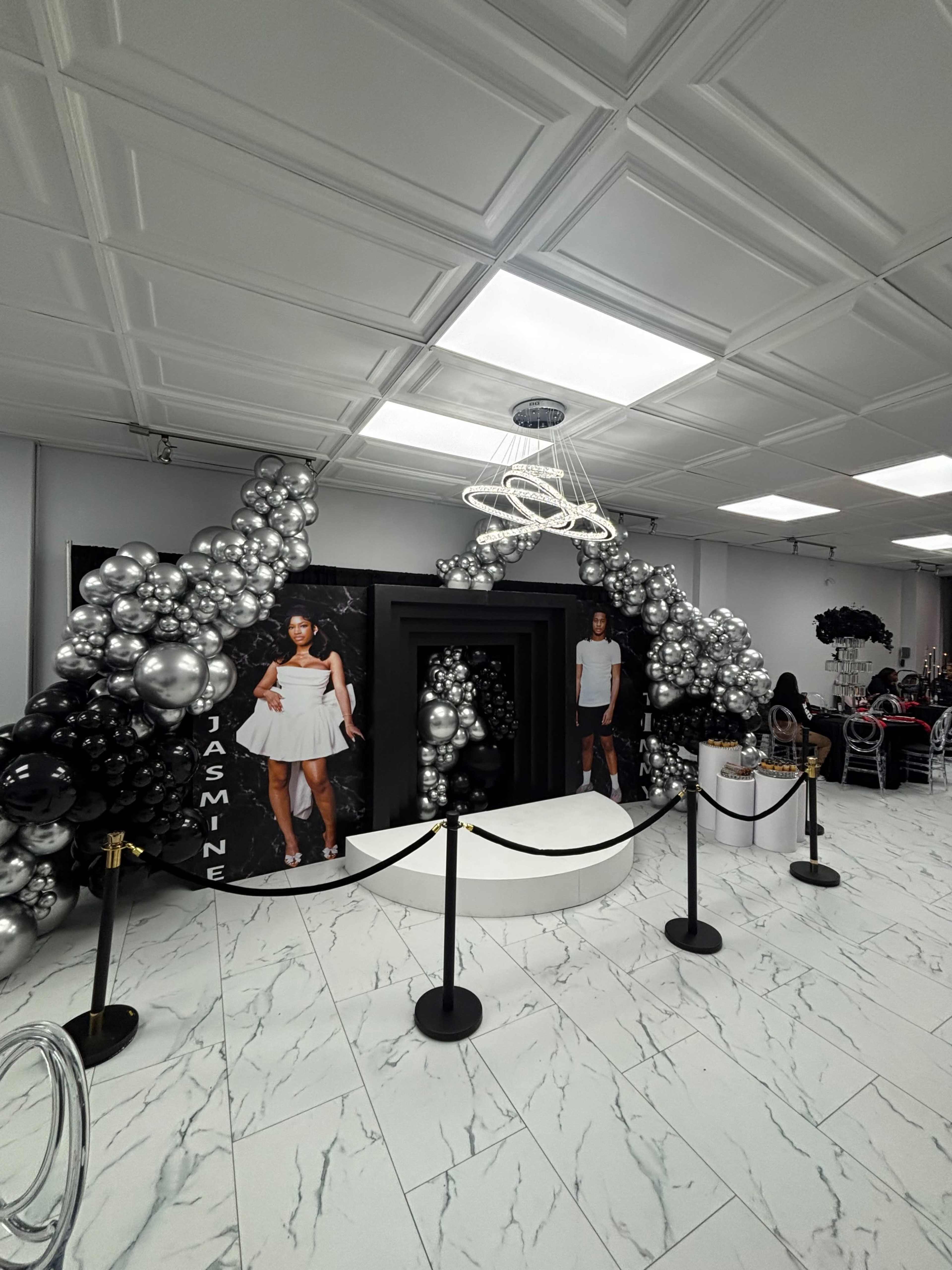 A decorated event space features a stylish balloon arch in black and silver, with a central white stage and two life-size cutouts of a woman in a white dress.
