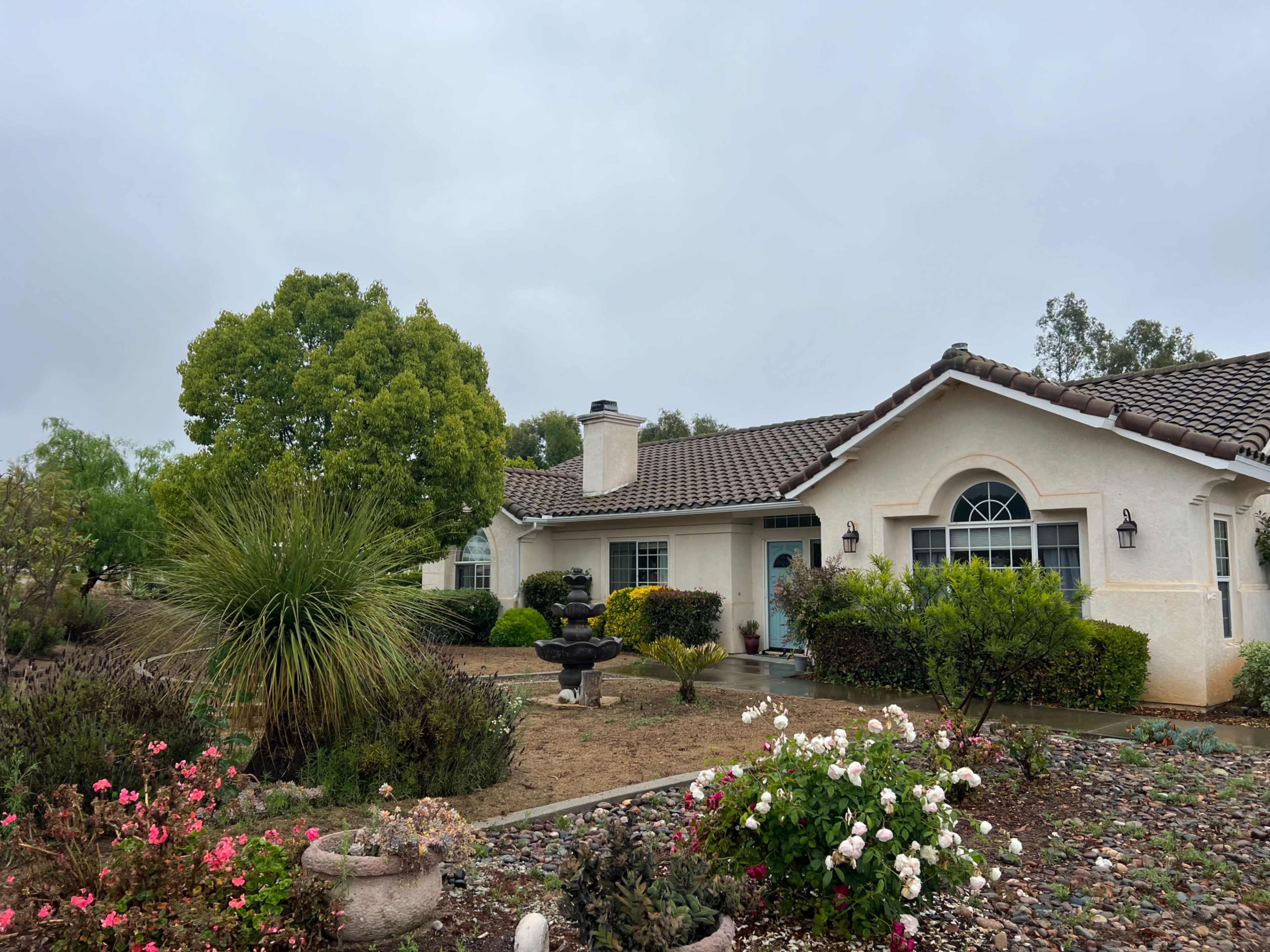 The image shows a single-story house with a tiled roof, surrounded by a landscaped yard featuring various shrubs and flower beds.