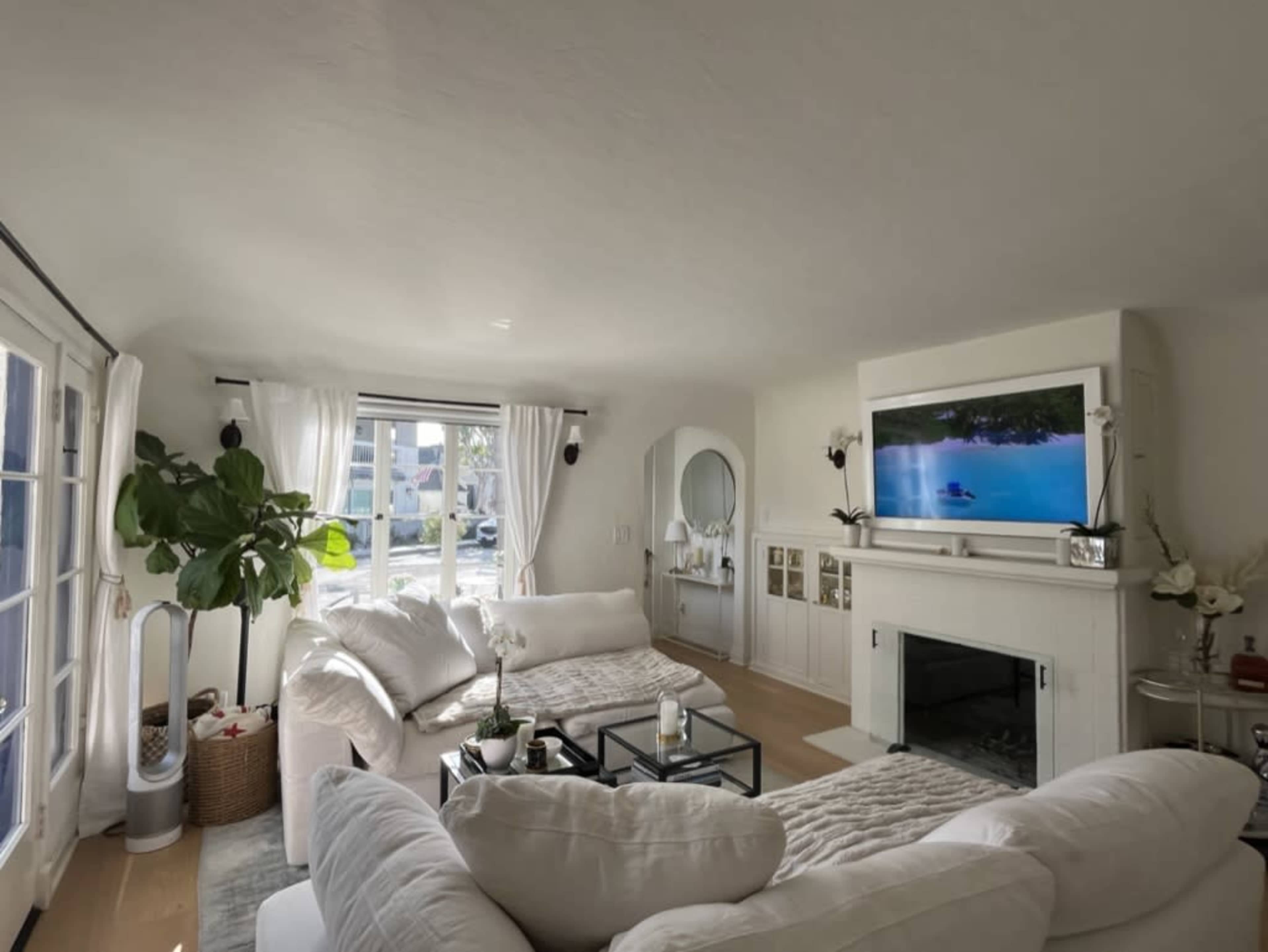 The living room features two large white sofas, a black coffee table, a flat-screen TV mounted above a white fireplace, and large windows with sheer curtains, allowing natural light to enter.