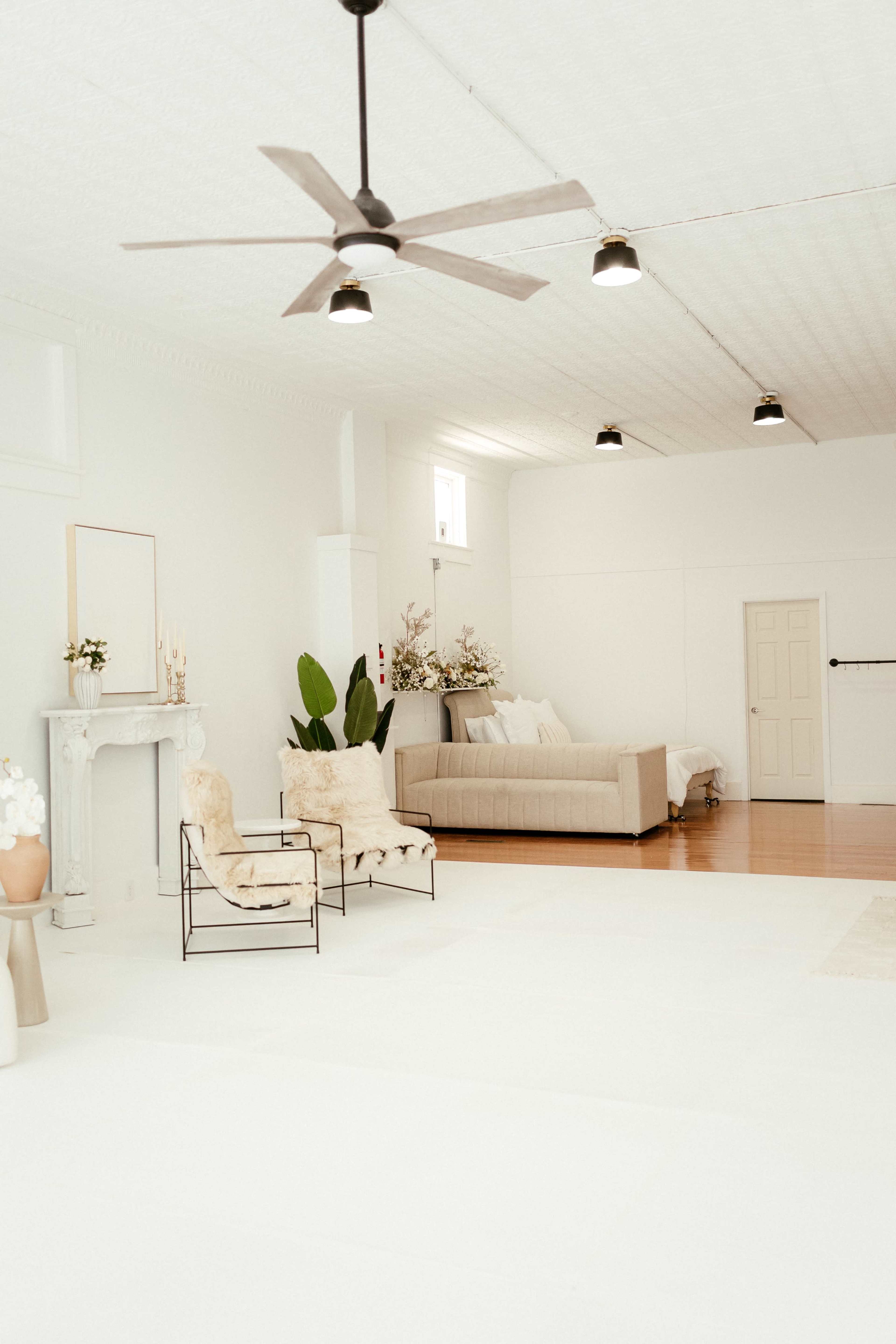 The image shows a spacious, well-lit room with white walls, a light-colored sofa, and minimal furniture including a pair of chairs, decorative plants, and a mantle.