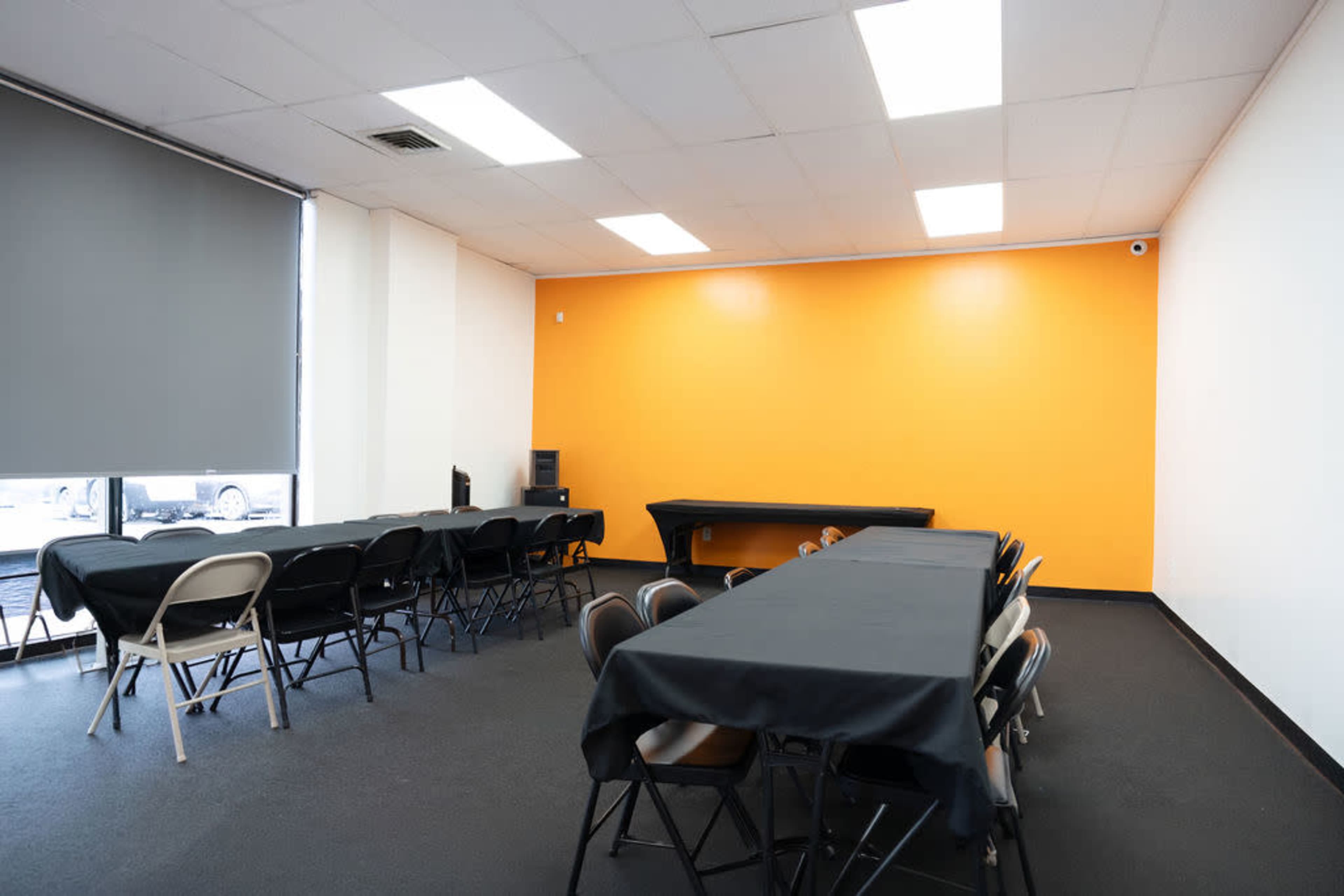 The image shows a simple, empty room with multiple black tables and chairs arranged neatly, highlighted by an orange accent wall.