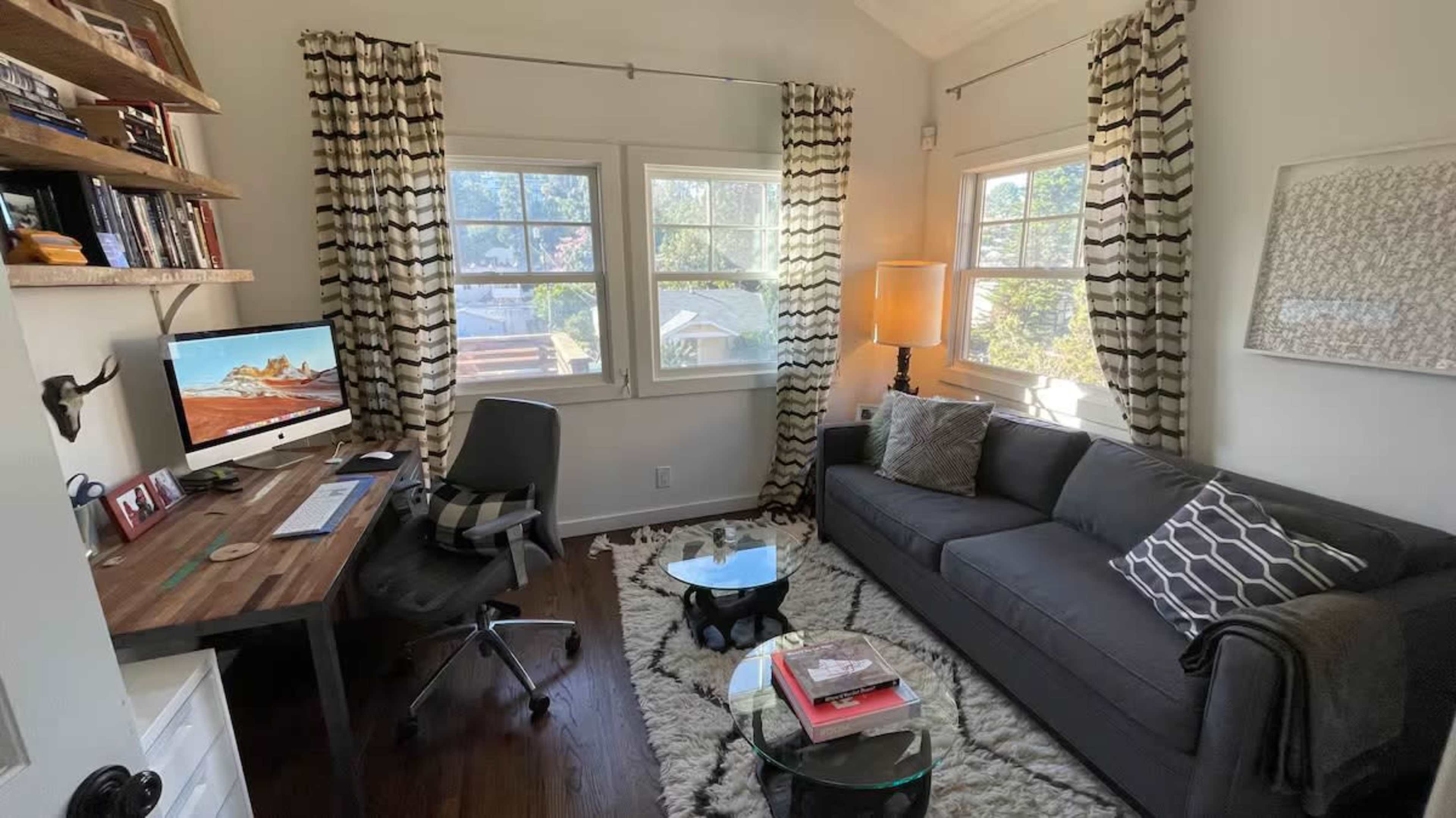 A cozy home office features a desk with a computer, a dark gray sofa, and large windows dressed with patterned curtains, all arranged on a shaggy area rug.