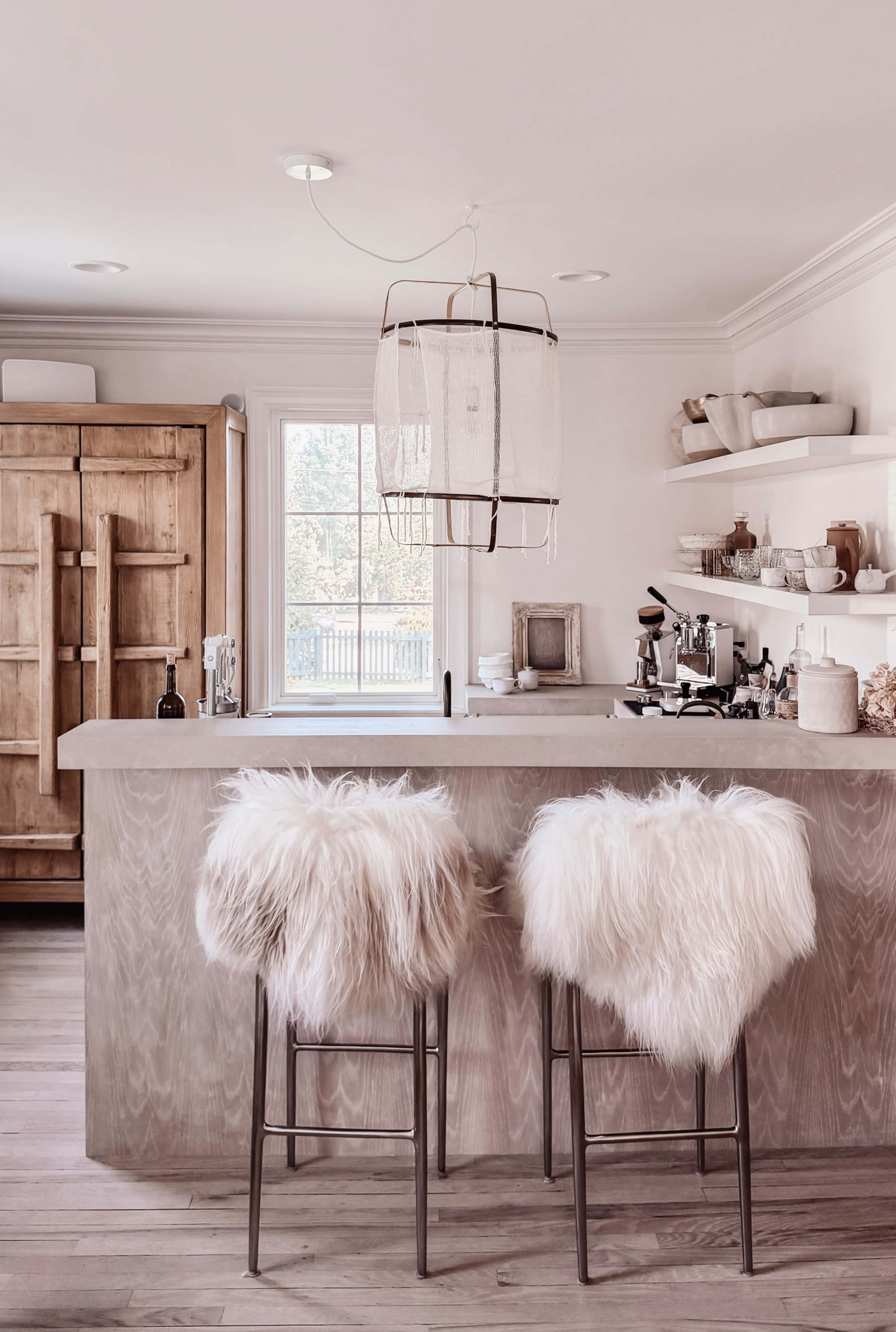 The image shows a modern kitchen with a wooden island, two bar stools covered in white fur, and a window allowing natural light to enter the space.