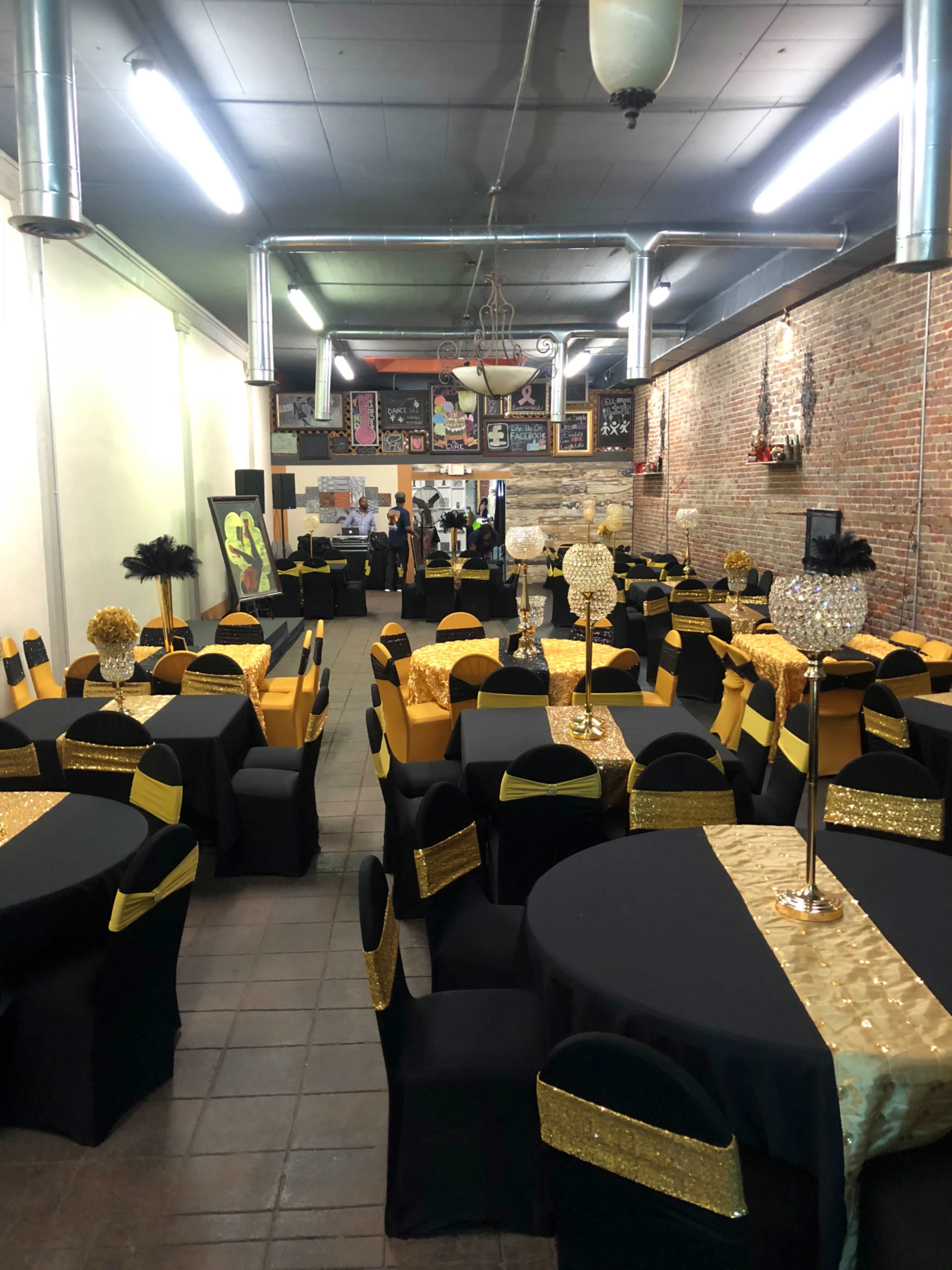 A large event space set up with black and gold tables and chairs, featuring decorative centerpieces and a brick wall in the background.