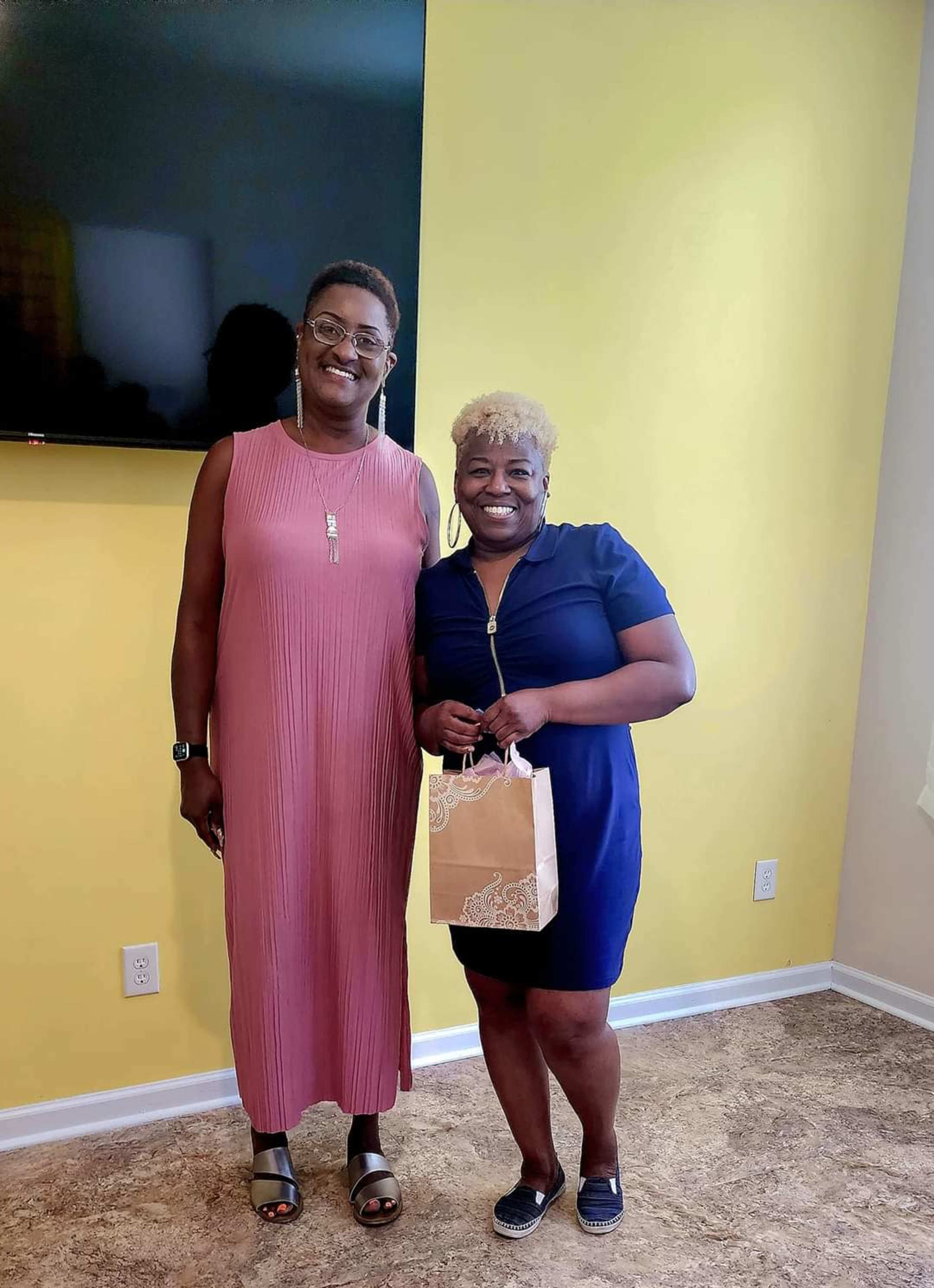Two women stand smiling in a brightly lit room, one wearing a long pink dress and the other in a dark blue dress, while holding a small gift bag.