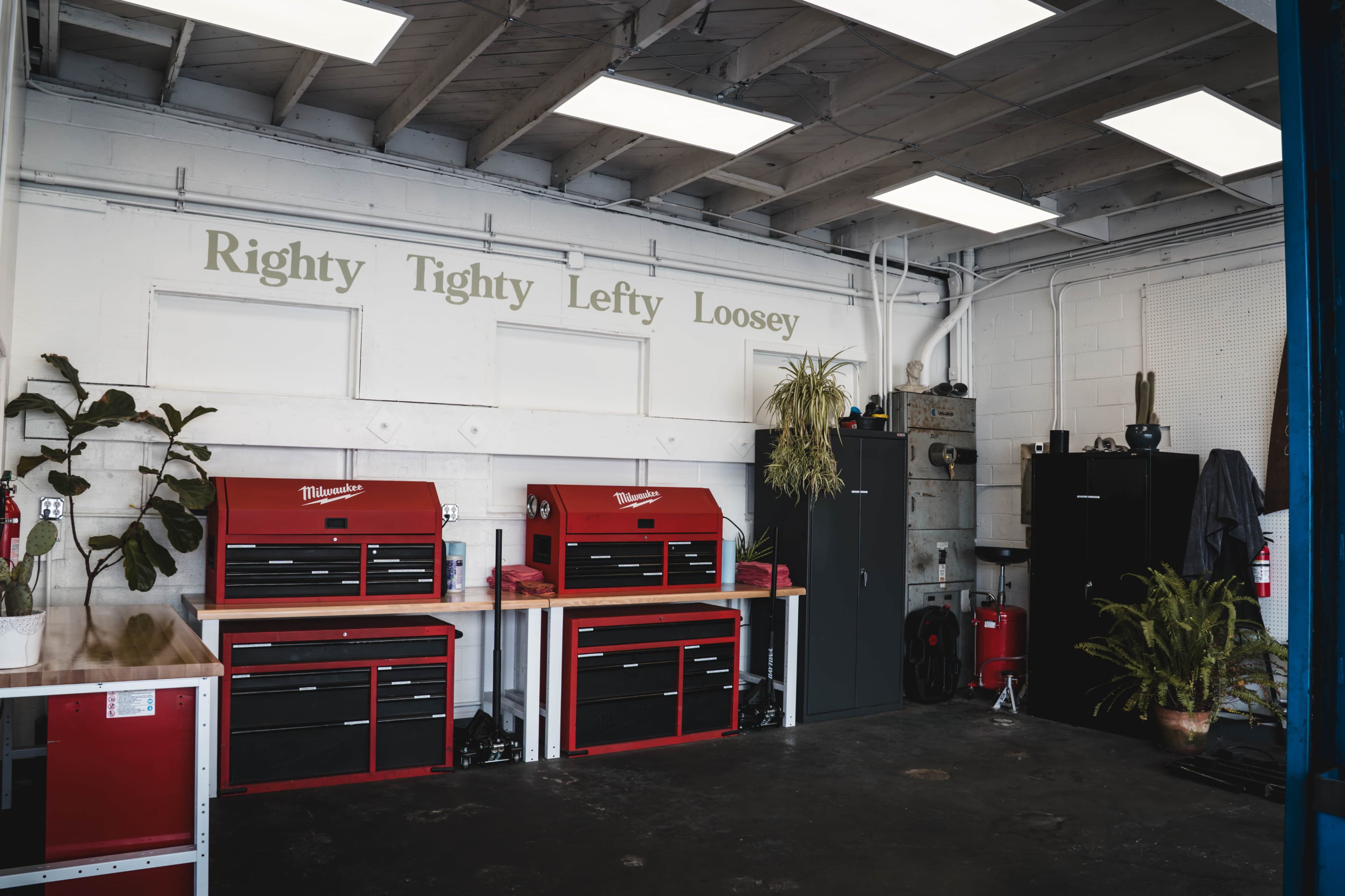 The image shows a garage or workshop featuring two red tool chests against a white wall that displays the phrase "Righty Tighty Lefty Loosey," with a potted plant beside the chests.