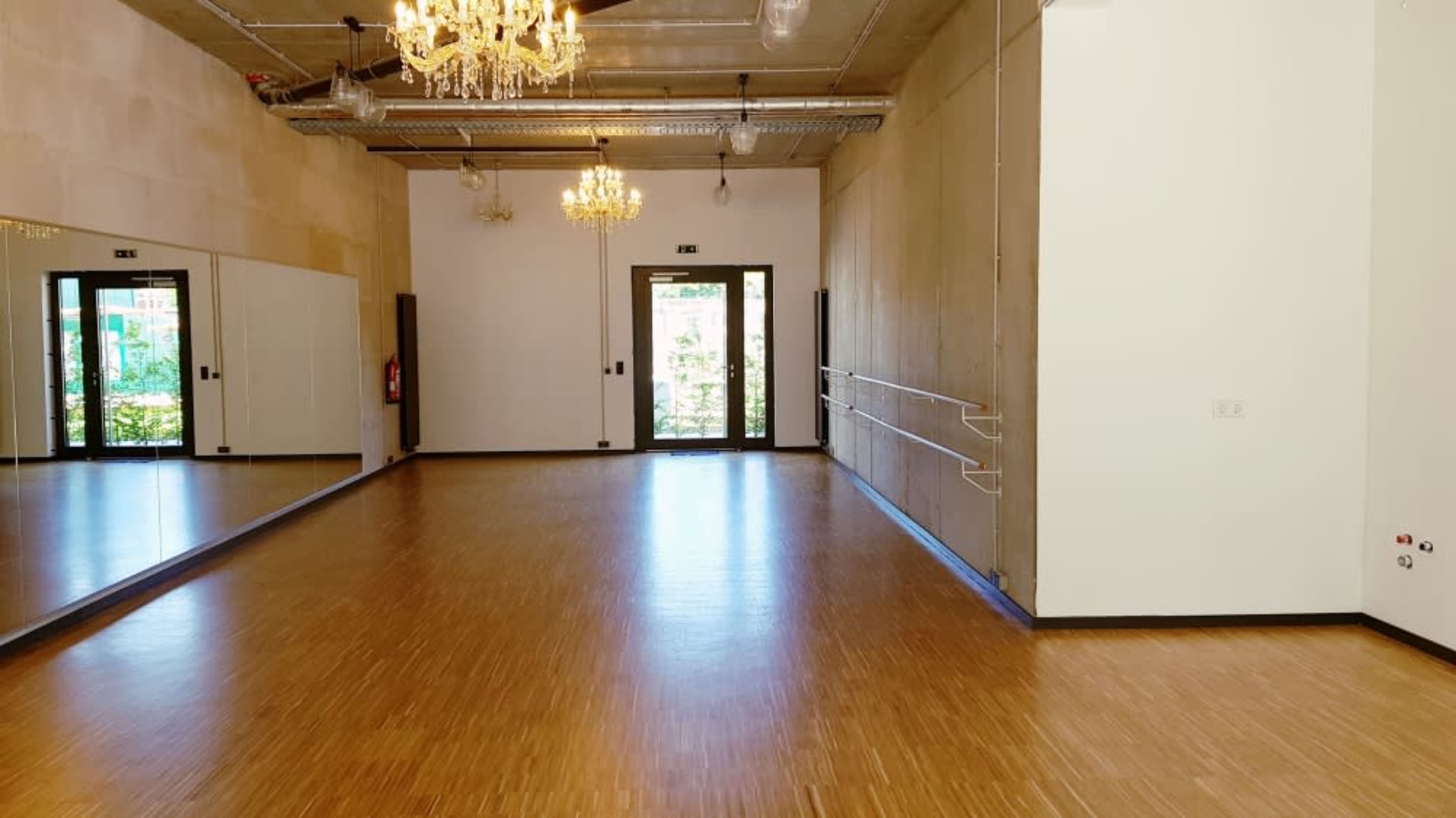 A spacious room with hardwood flooring, large mirrors on one wall, and two chandeliers hanging from the ceiling.