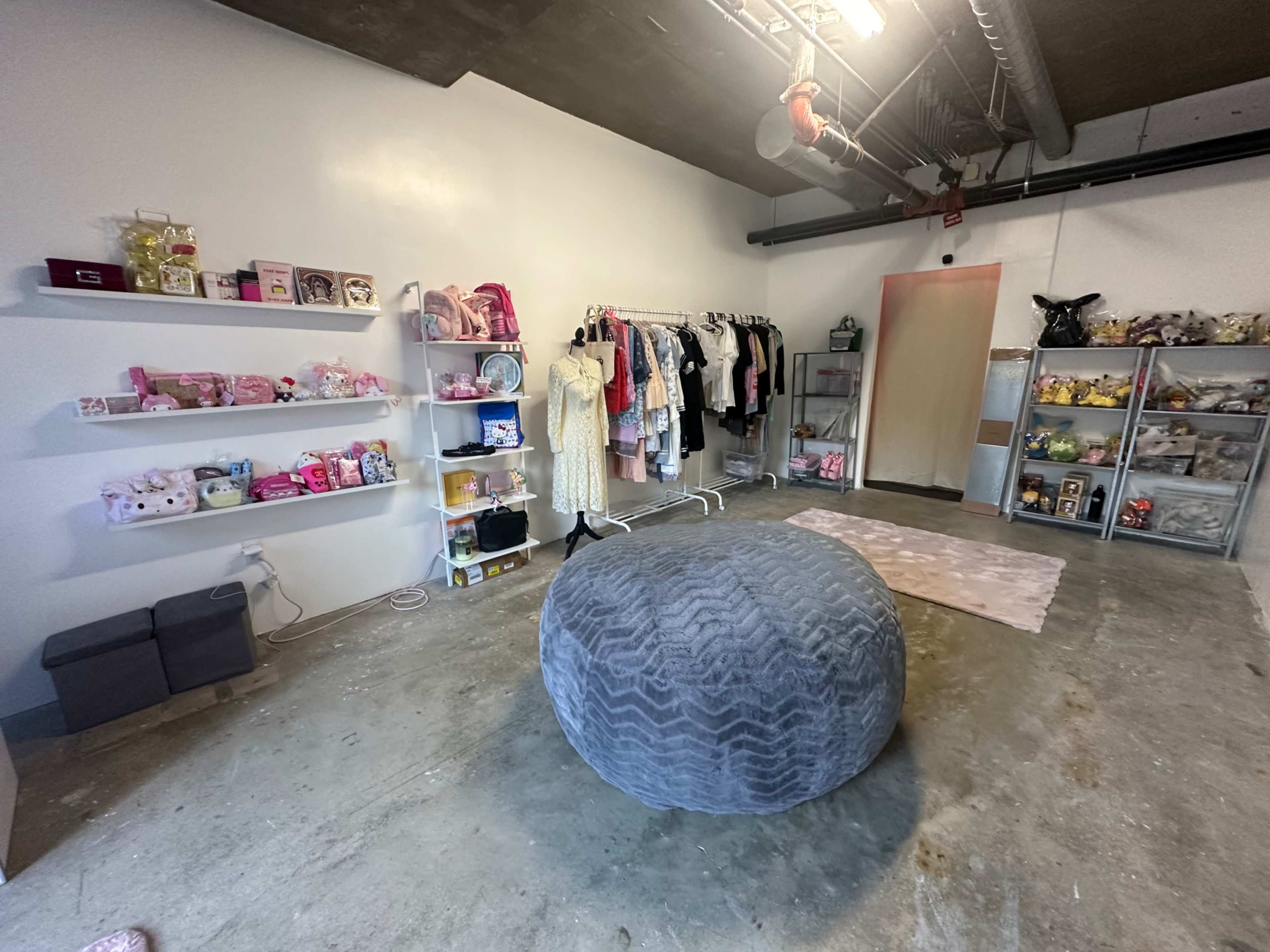 The room features a large gray pouf, clothing racks, shelves with various items, and a light-colored rug on the concrete floor.
