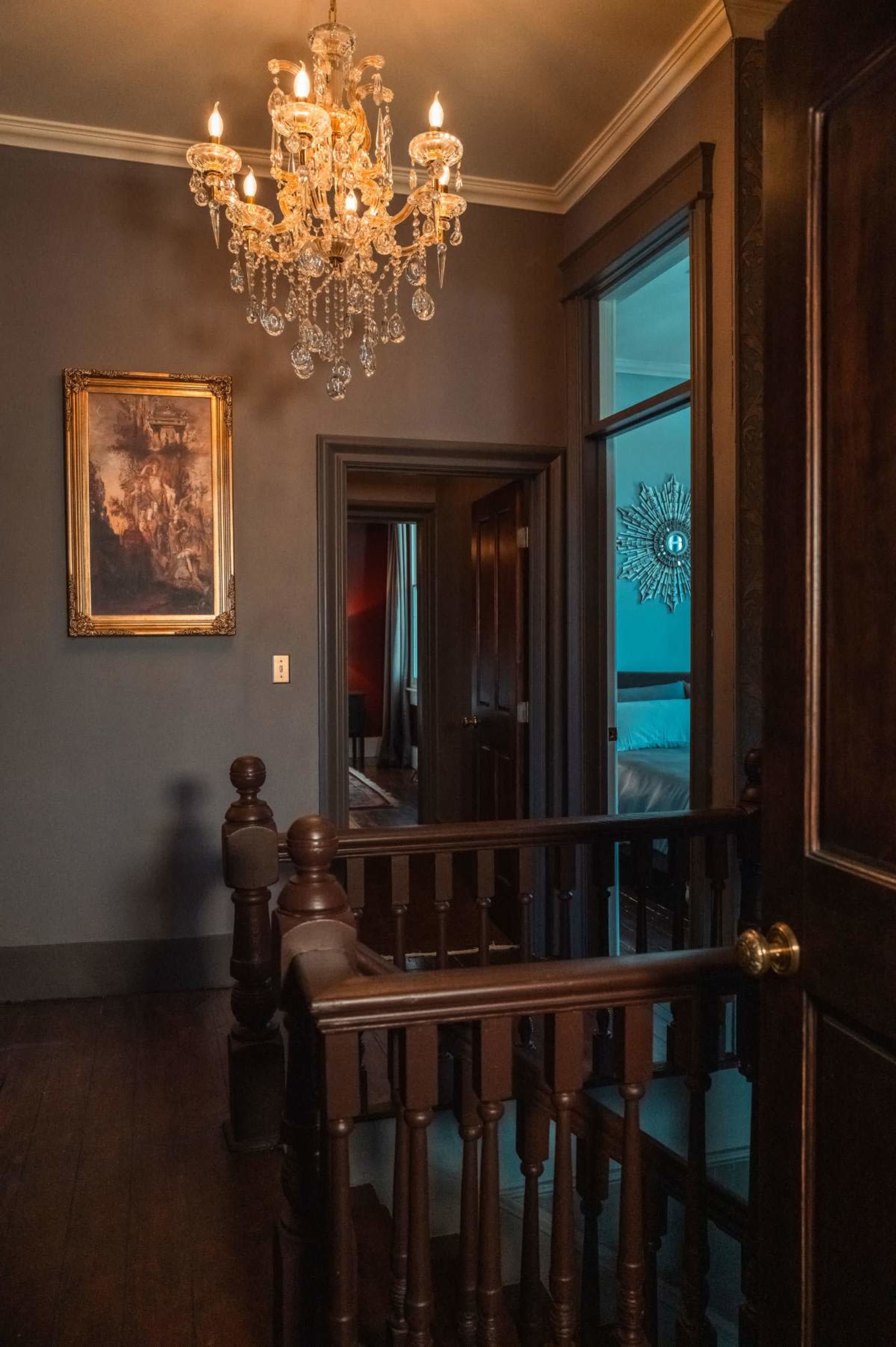 A wooden staircase leads to a dark hallway adorned with a chandelier and a framed painting on the wall.