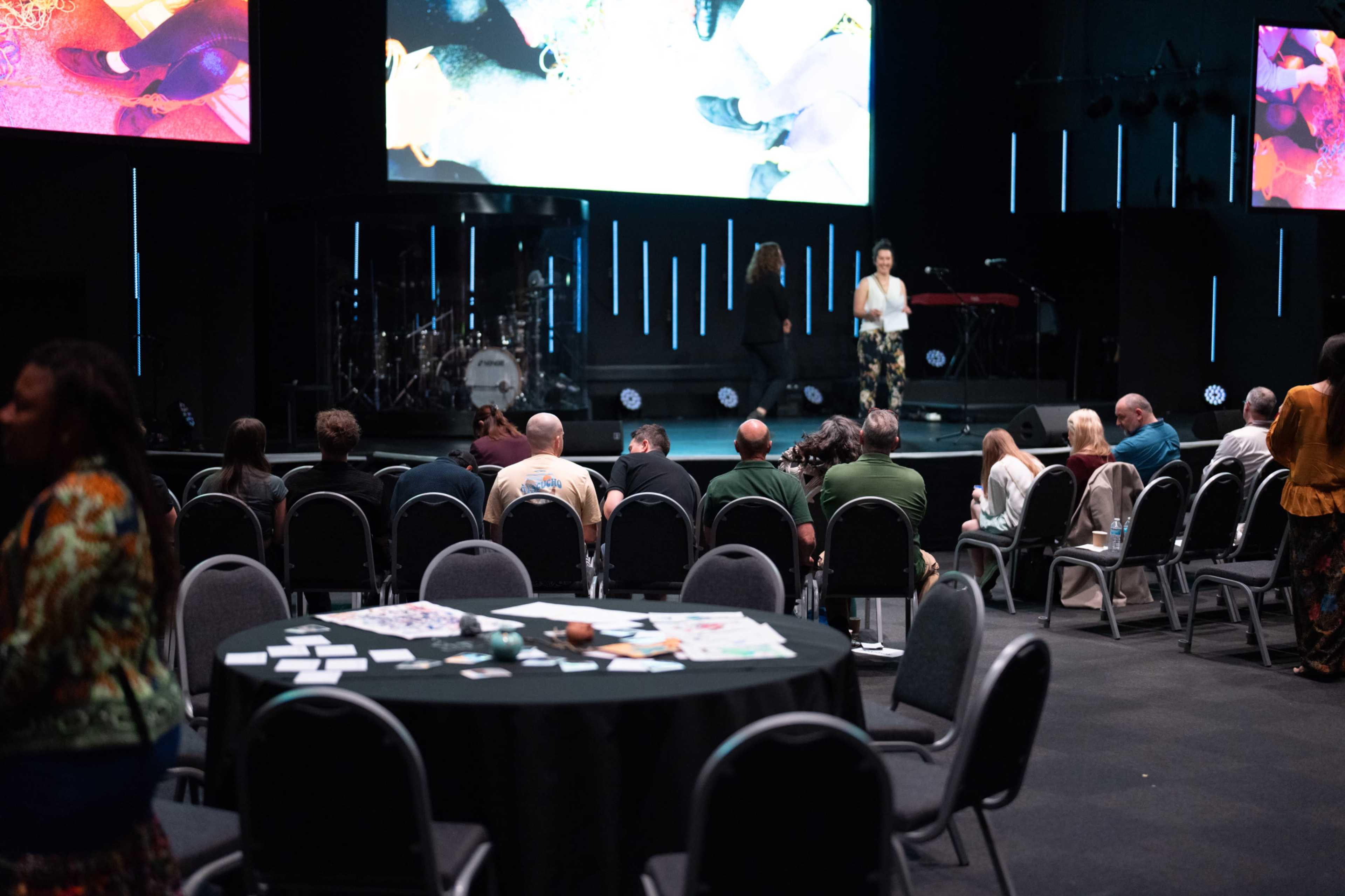 A speaker stands on a stage in front of a large screen while an audience sits in chairs arranged in rows in a venue.