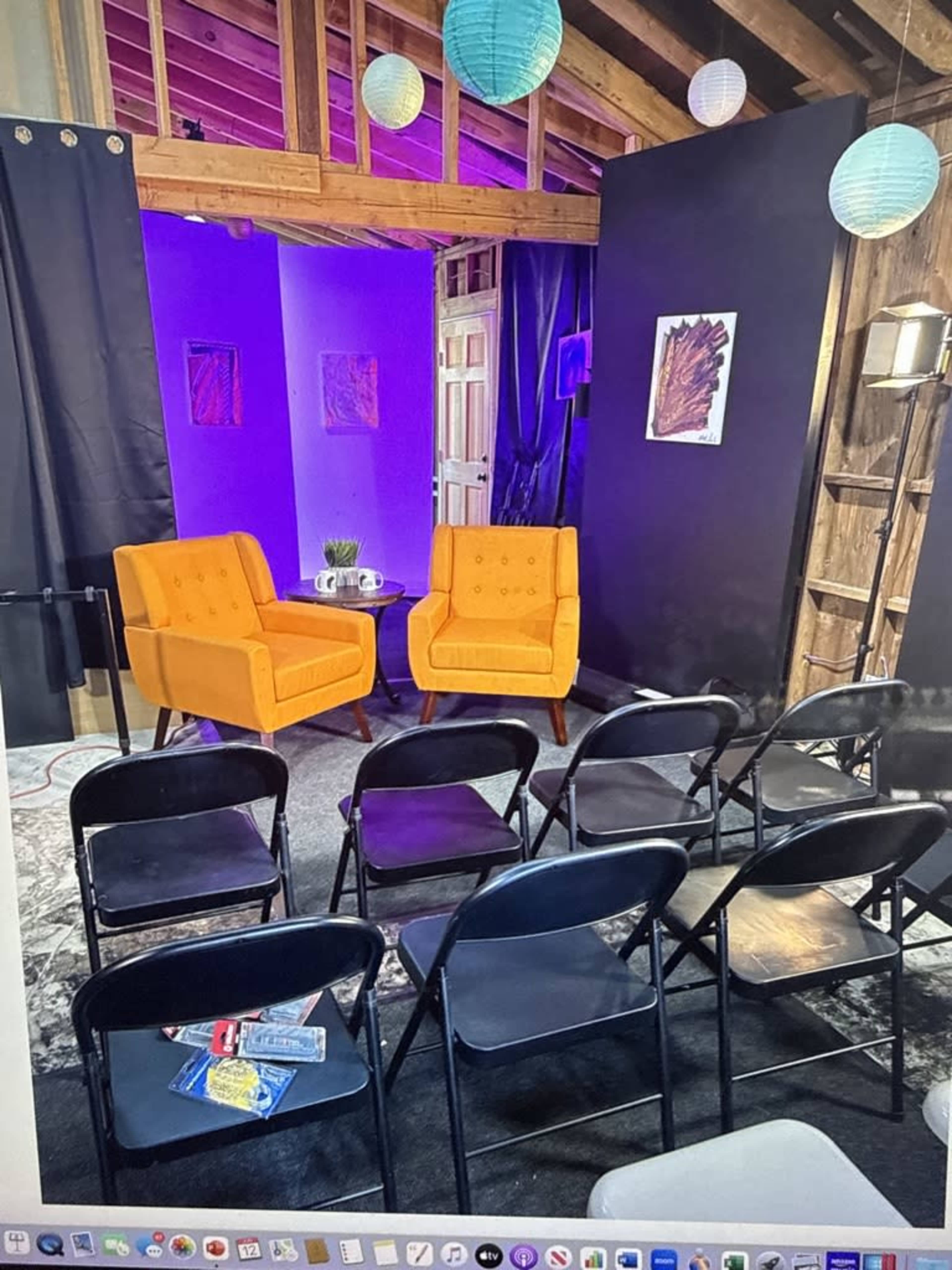 The image shows a cozy stage setup featuring two orange chairs facing a small table, with black folding chairs arranged in front and colorful lanterns hanging from the ceiling.