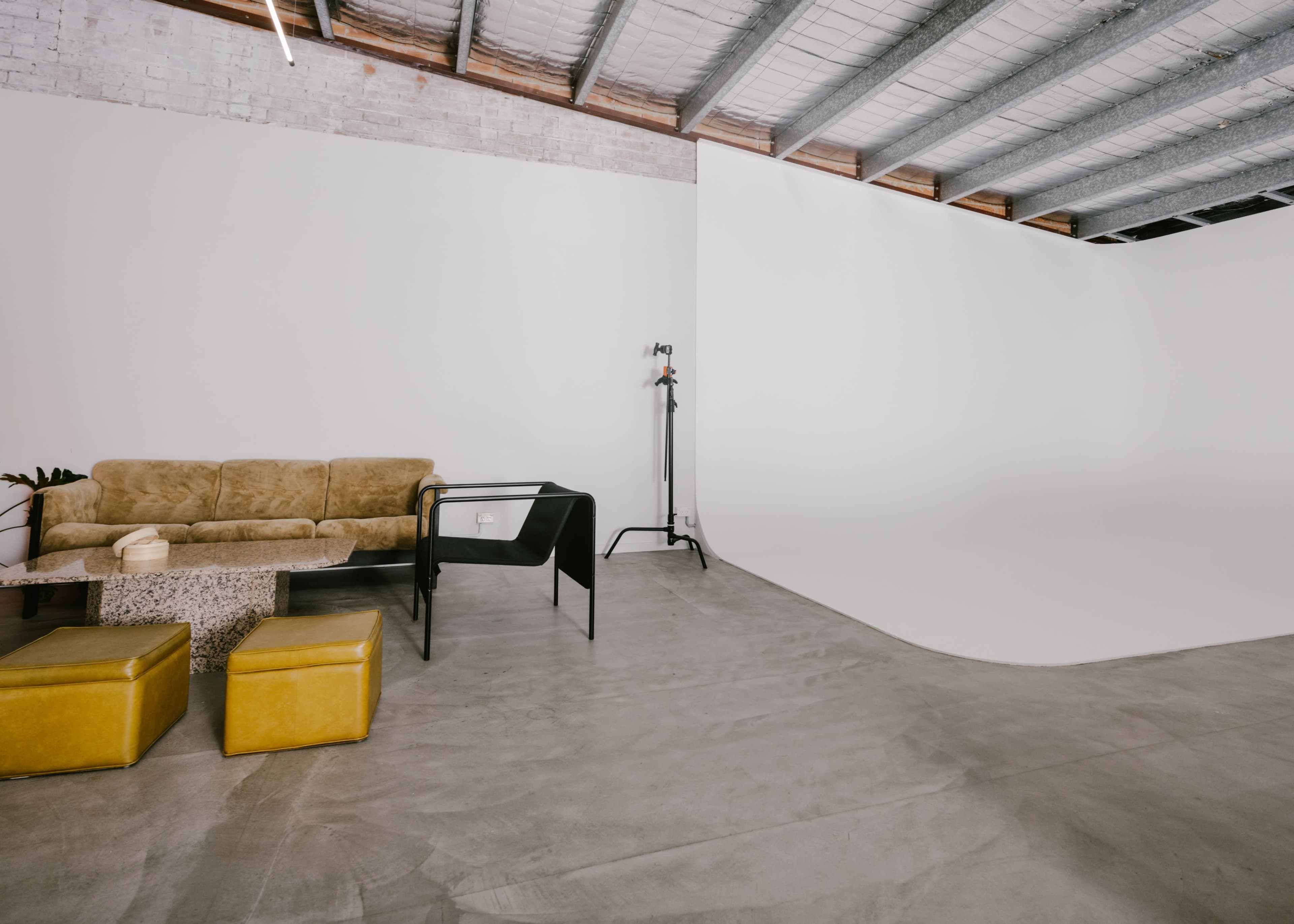 A minimalist studio space featuring a brown couch, a glass-top table, yellow ottomans, and a backdrop for photography.