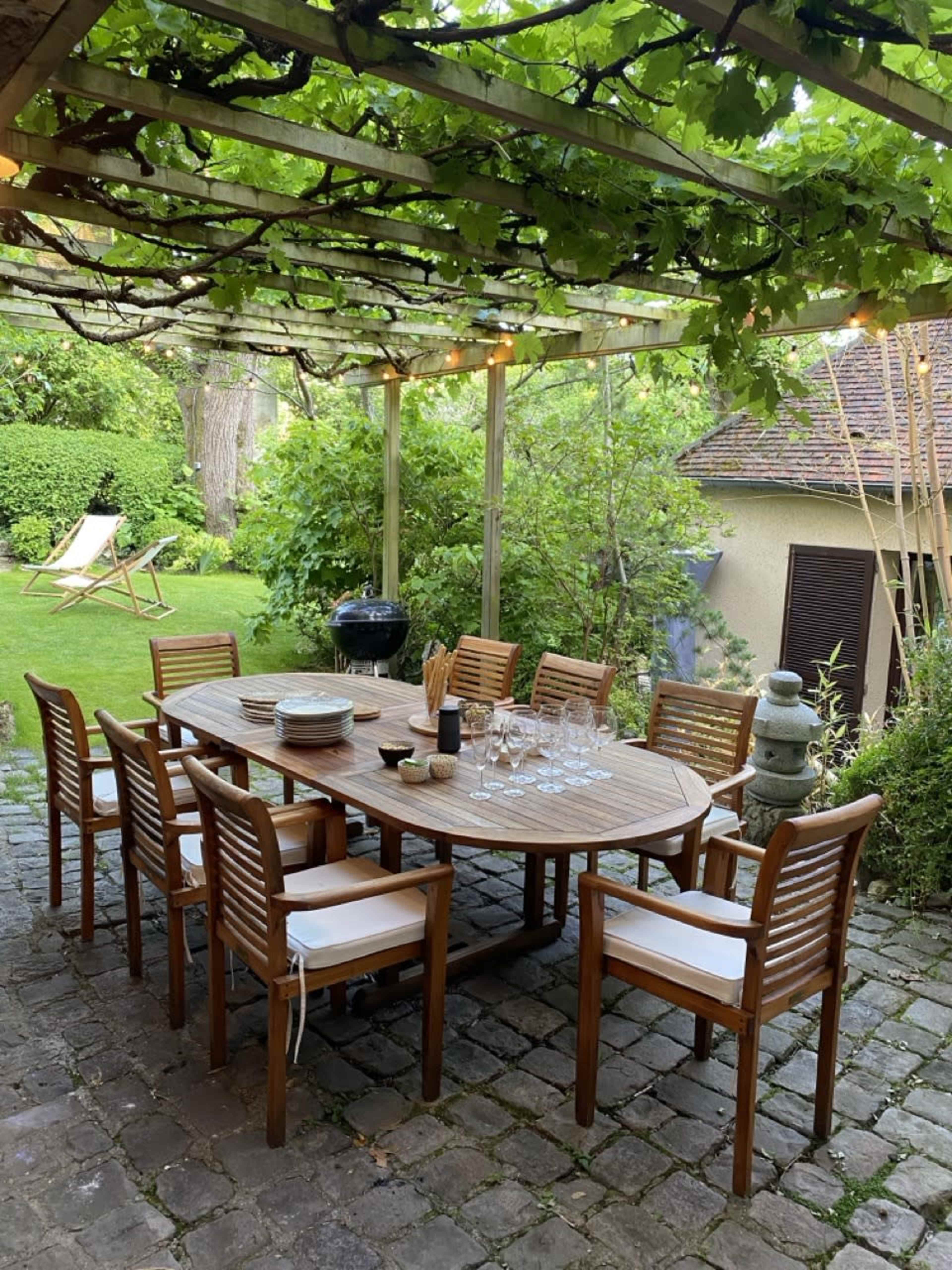 A wooden dining table is set with dishes and glasses under a trellised pergola covered in vines, surrounded by a landscaped garden.