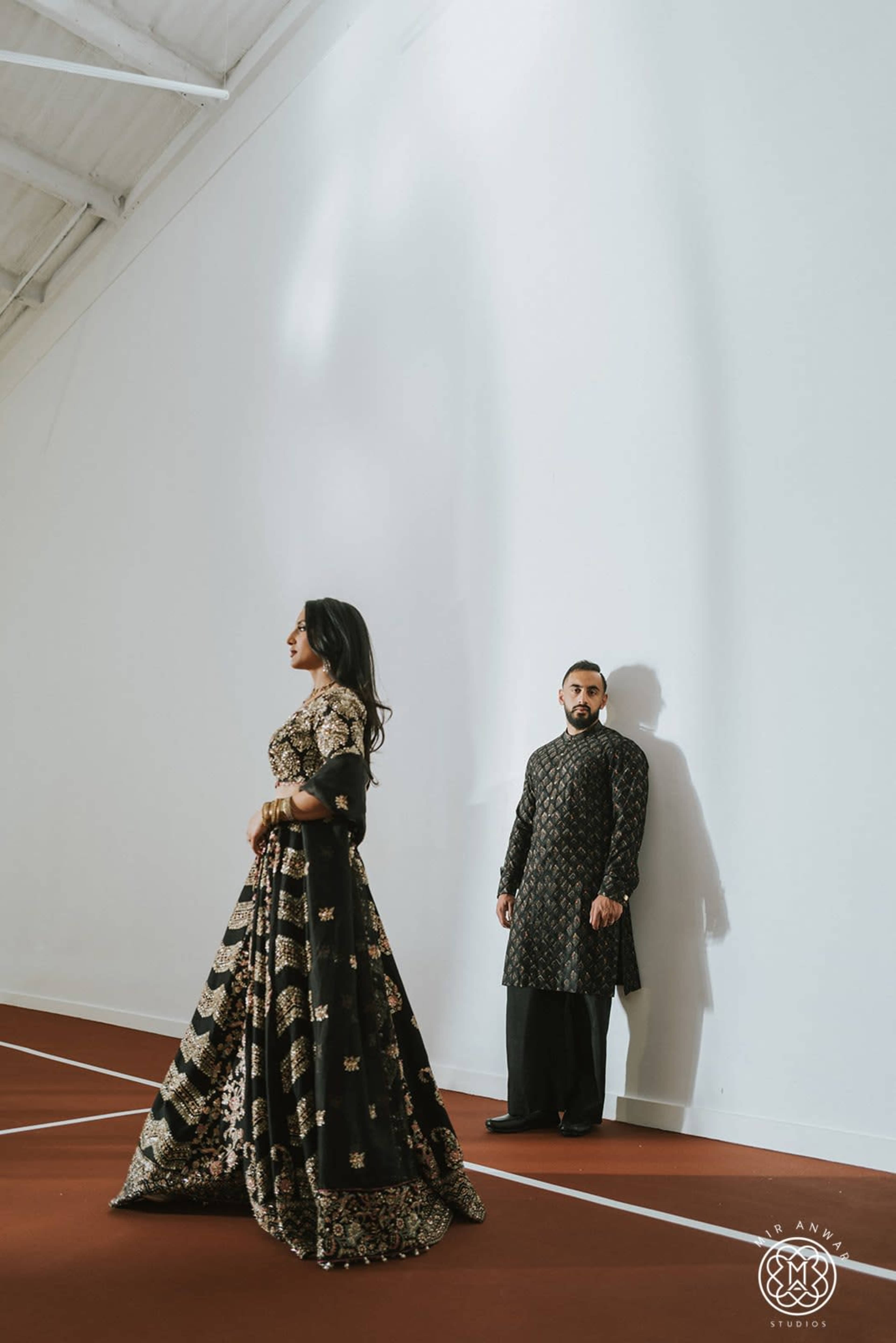 A man and a woman pose in an indoor space with a white wall, dressed in ornate traditional attire with intricate patterns.