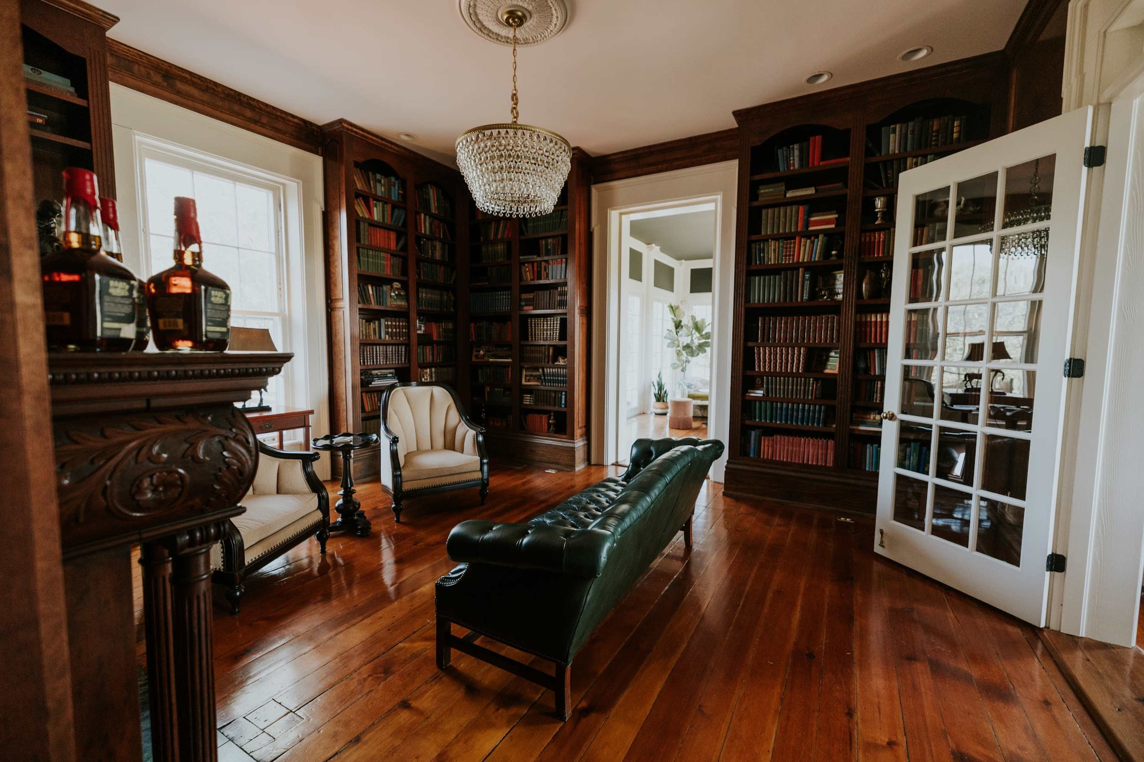 The image shows a cozy library with dark wooden bookshelves lined with books, a green leather chaise lounge, and a small round table next to a plush armchair, all under a chandelier.