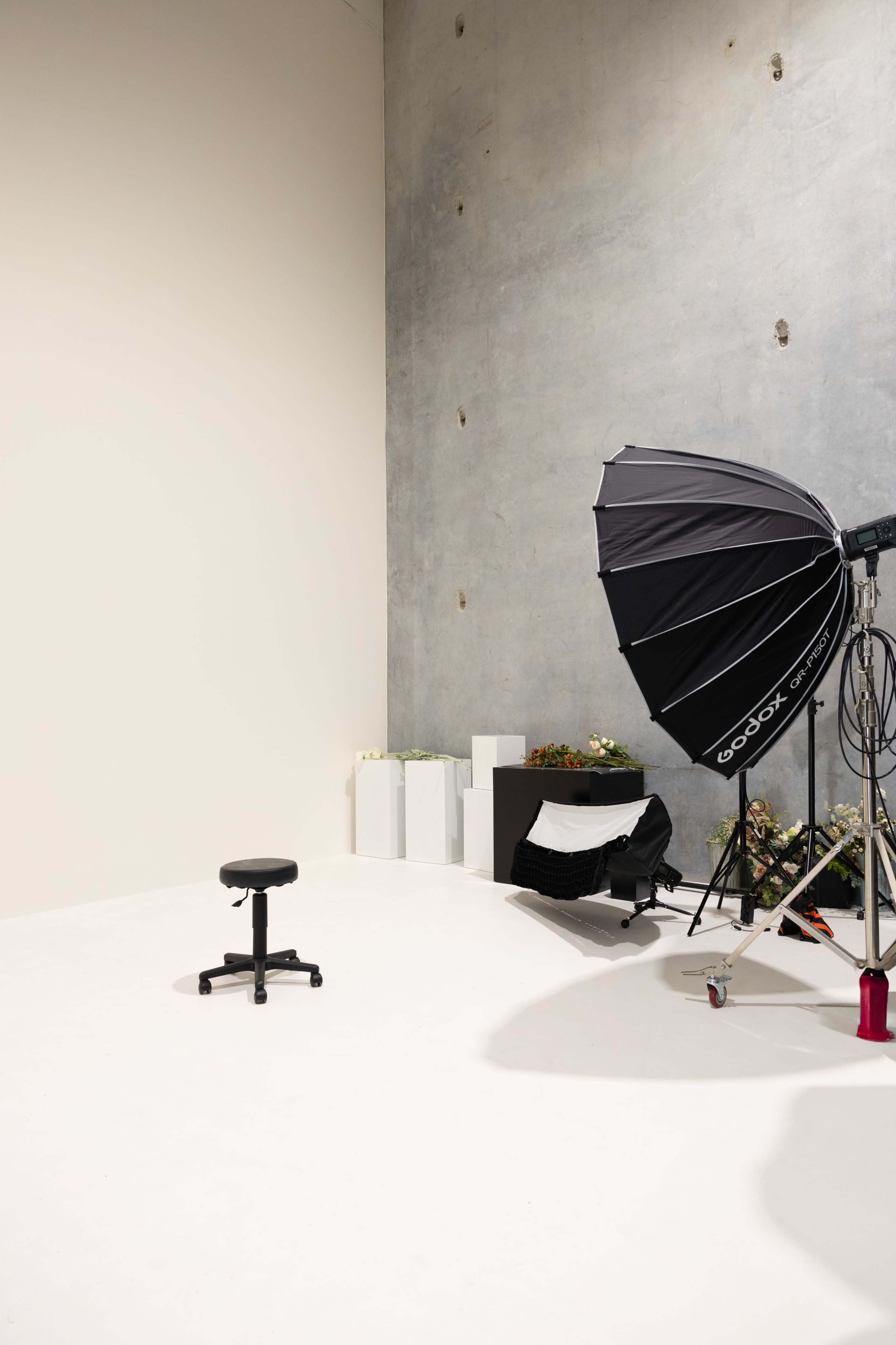 The image shows a photography studio with a black umbrella light, a stool, and minimalistic props against a plain wall.
