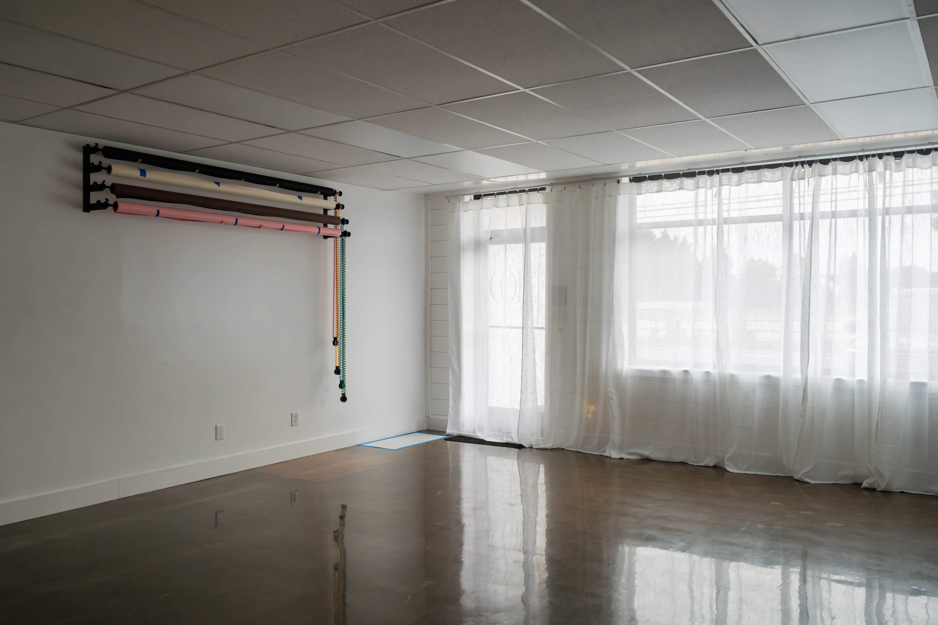 The image shows a spacious, empty room with a polished floor, large windows draped with sheer curtains, and a wall-mounted rack containing rolls of colored material.