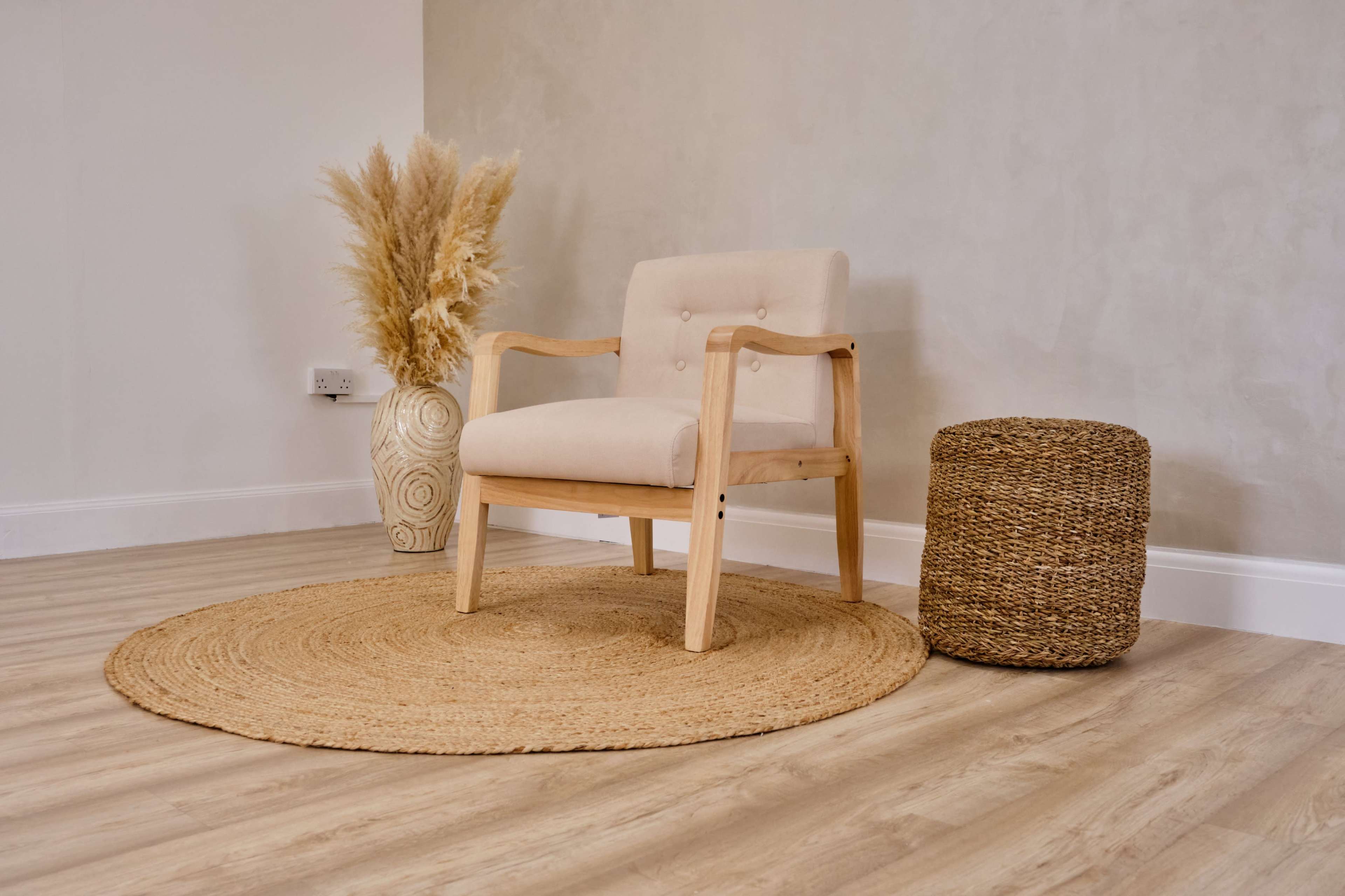 A minimalistic interior setting featuring a light wood chair, a round woven rug, a wicker side table, and a decorative vase with dried plants.