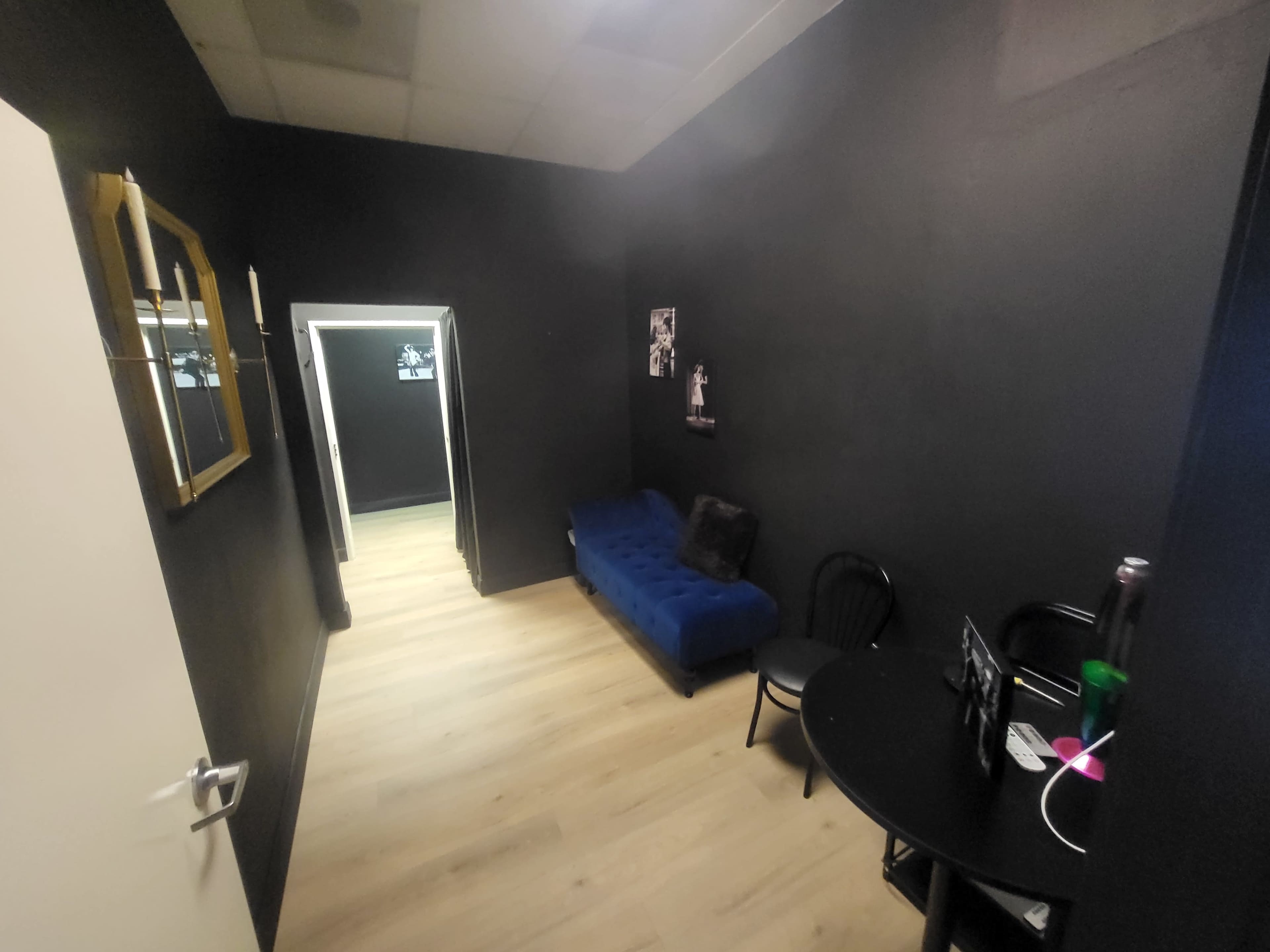 The image shows a small, dimly lit room with black walls, a blue sofa, a round table with chairs, and an entrance to another space in the background.