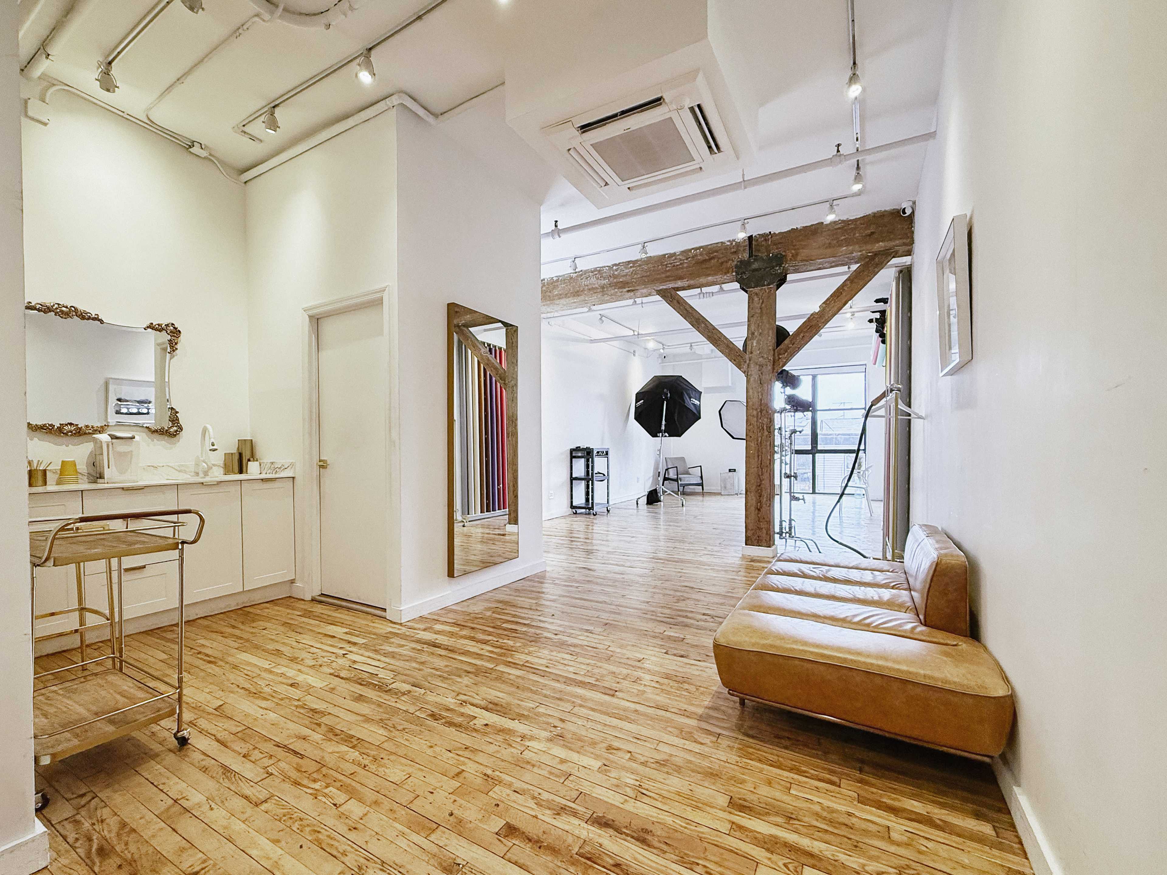 The image shows a spacious, well-lit interior with wood flooring, featuring a sofa, a large mirror, and photography equipment in the background.