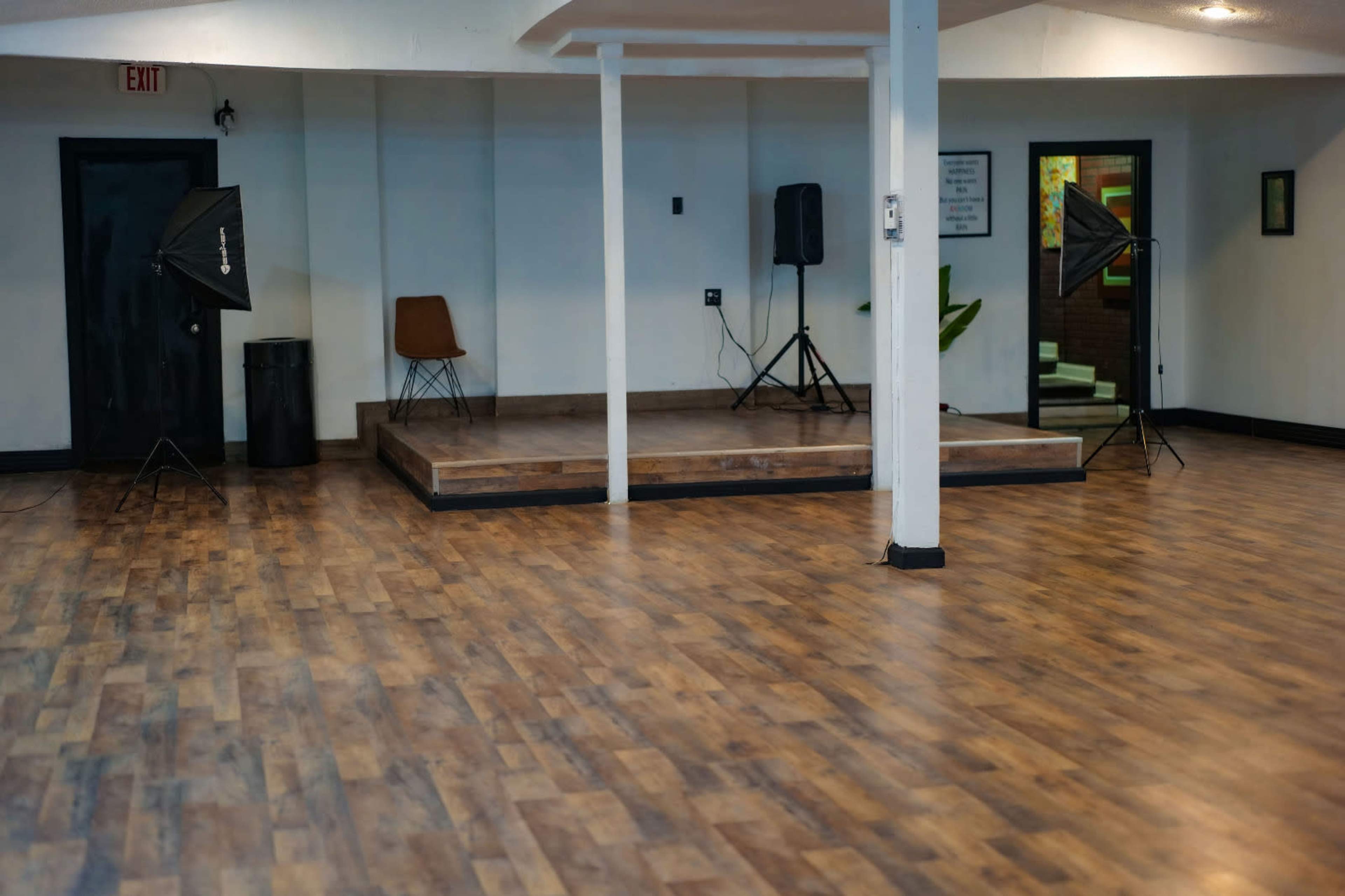 The image shows an empty indoor space with wooden flooring, a small stage, and photography equipment set up on either side.