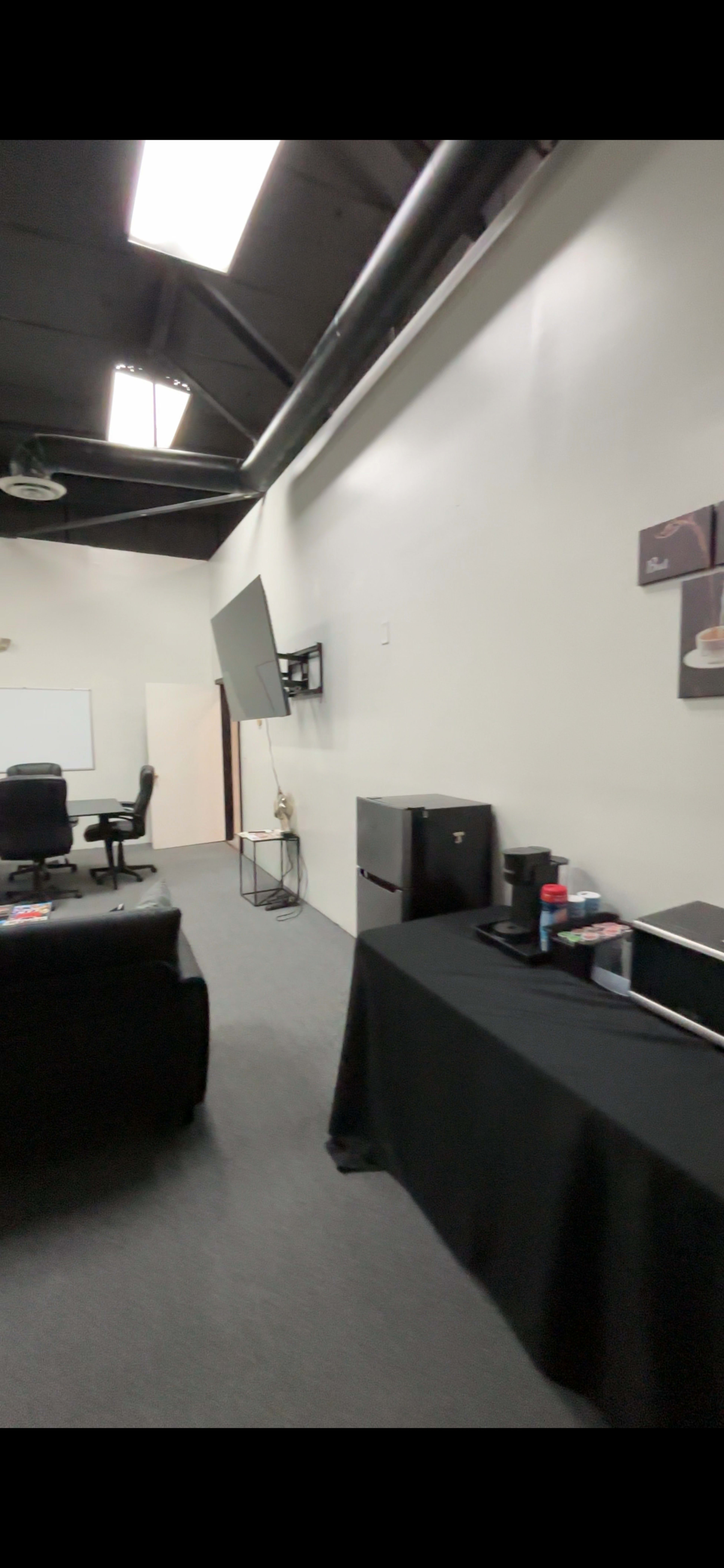 The image shows a modern office space with a black table displaying a coffee maker and supplies, a rolling chair, and a wall-mounted monitor, along with a whiteboard in the background.