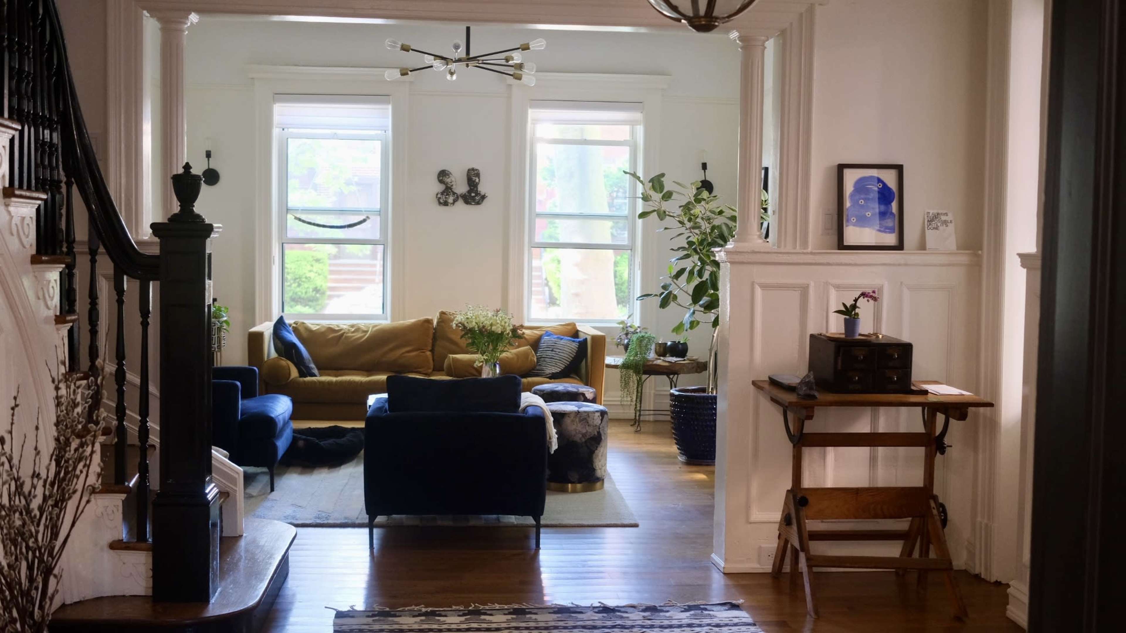 A well-lit living room with a mix of modern and vintage furniture, featuring a yellow sofa, dark blue armchairs, and houseplants, viewed from a hallway with a staircase.
