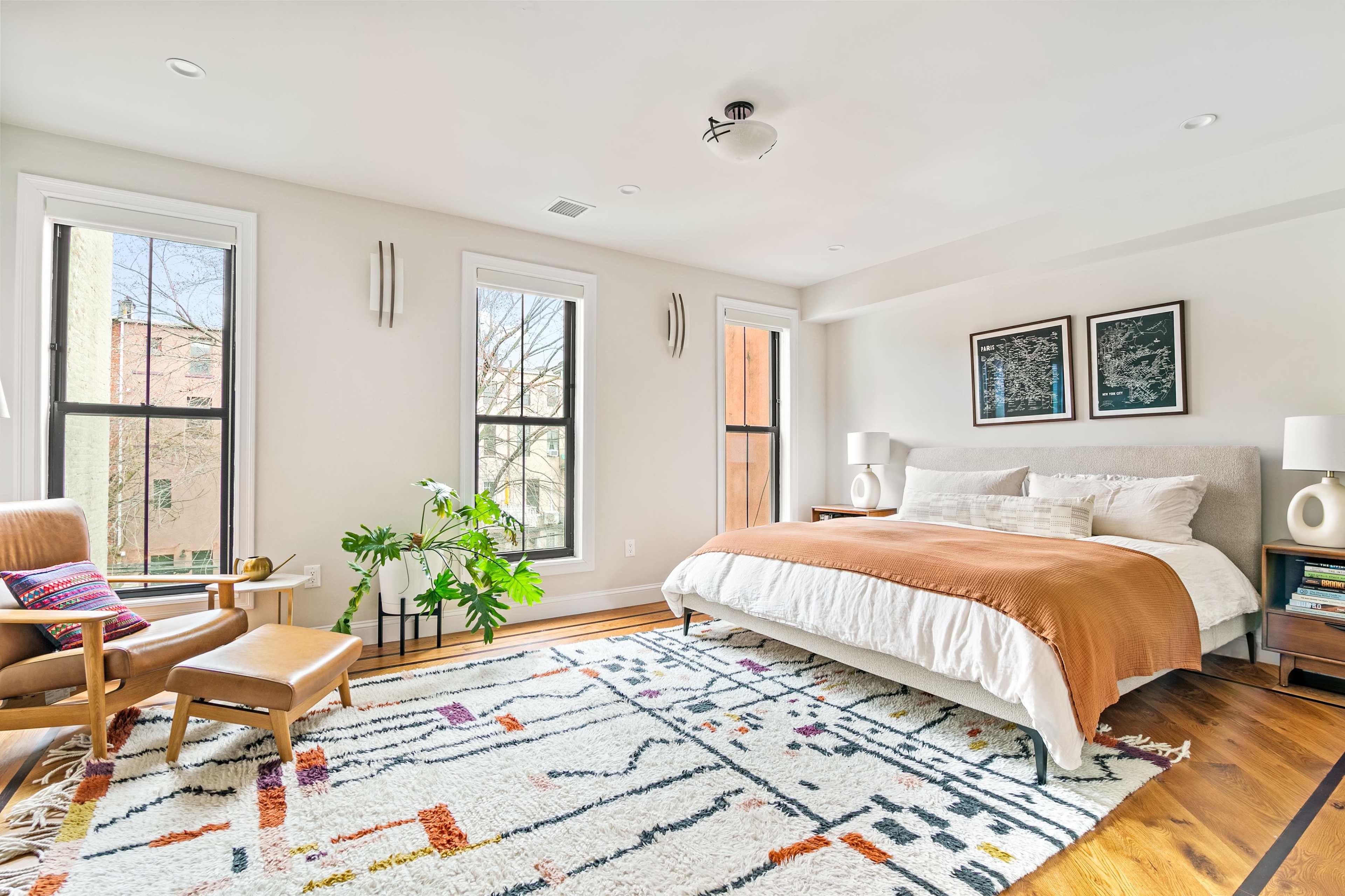 The image shows a bright bedroom with a large bed, two windows, a green plant, and a cozy armchair on a patterned rug.