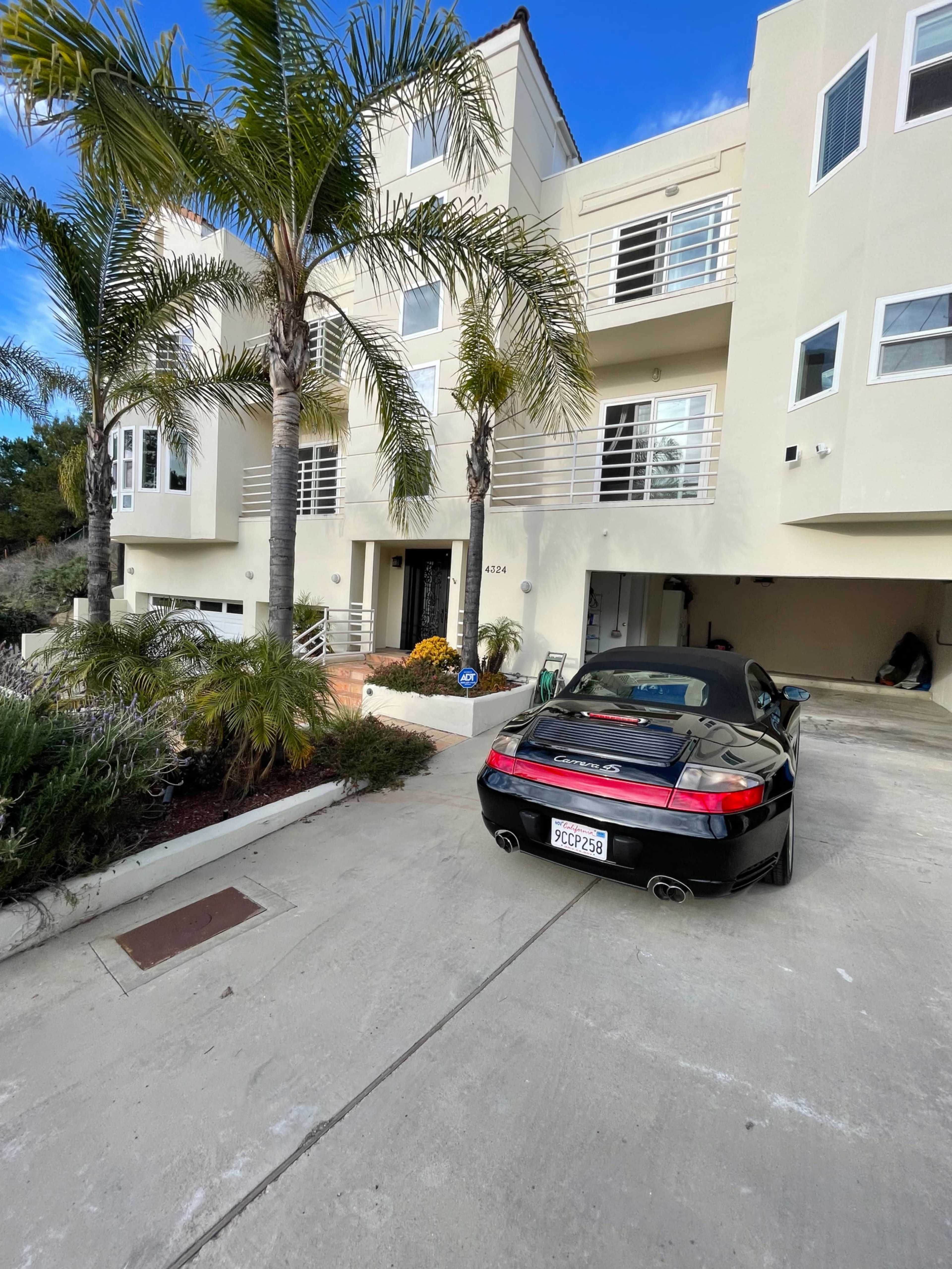 A black sports car is parked in the driveway of a modern, two-story house surrounded by palm trees.