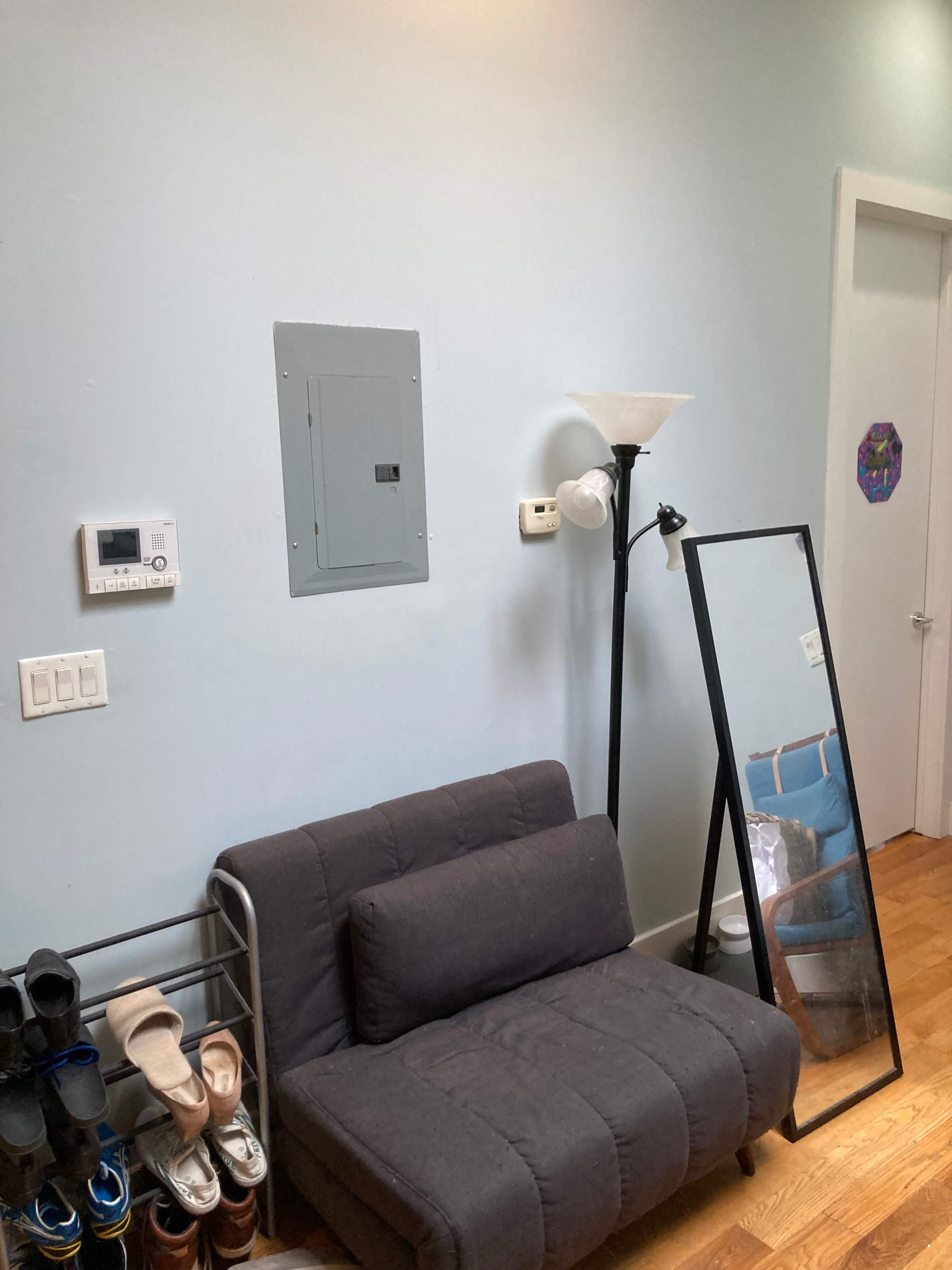 A small living space featuring a gray sofa, a shoe rack, a floor lamp, a full-length mirror, and an electrical panel on a light blue wall.