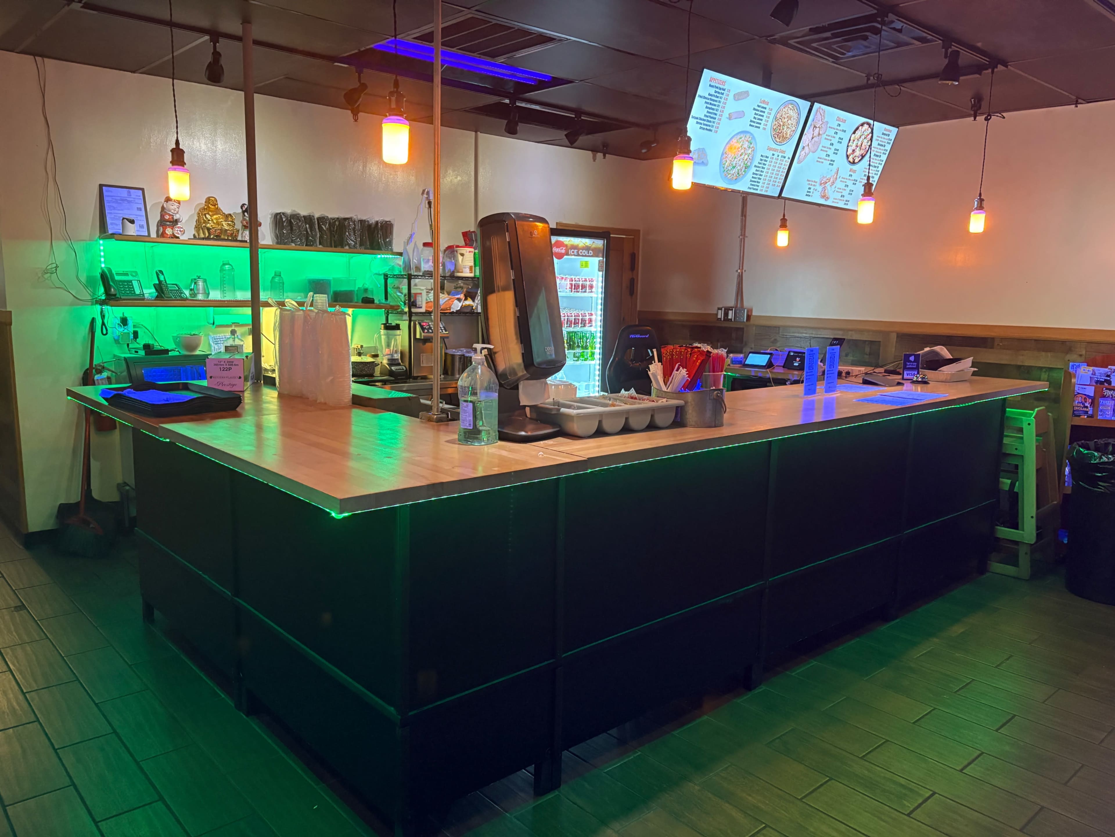 The image shows a modern restaurant counter with a long wooden surface, various drink dispensers, and illuminated menu boards above.