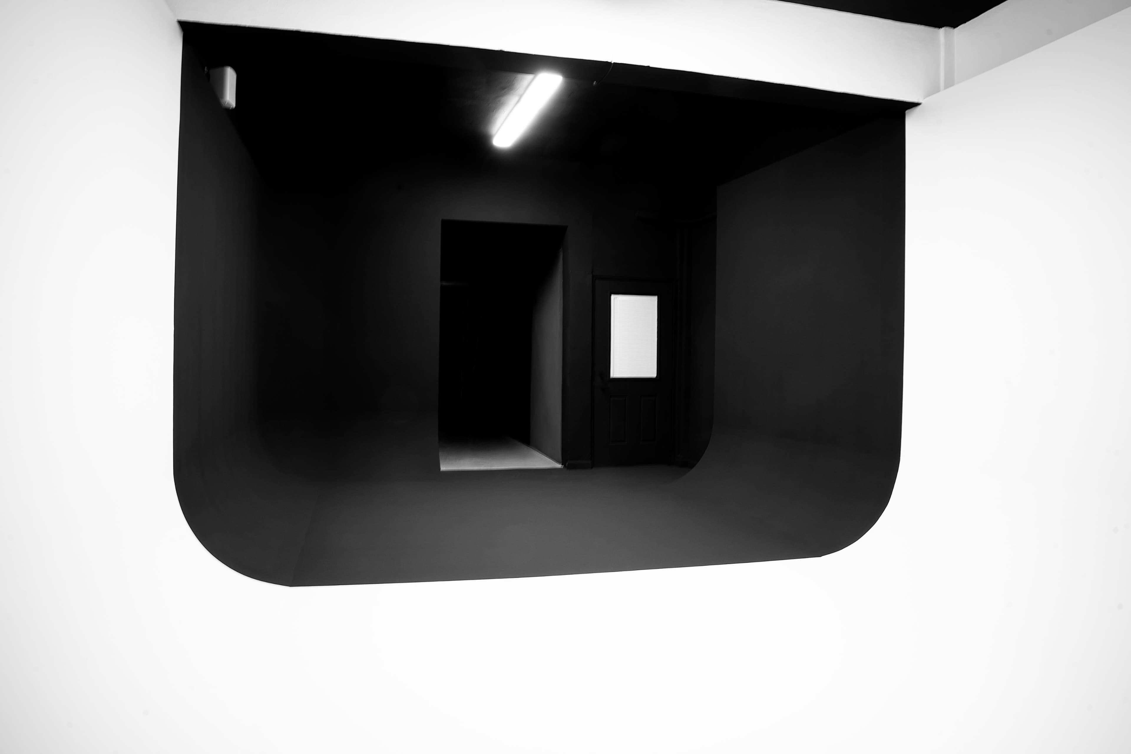The image shows a black and white interior space with a large, rounded opening in a white wall leading to a dark doorway.