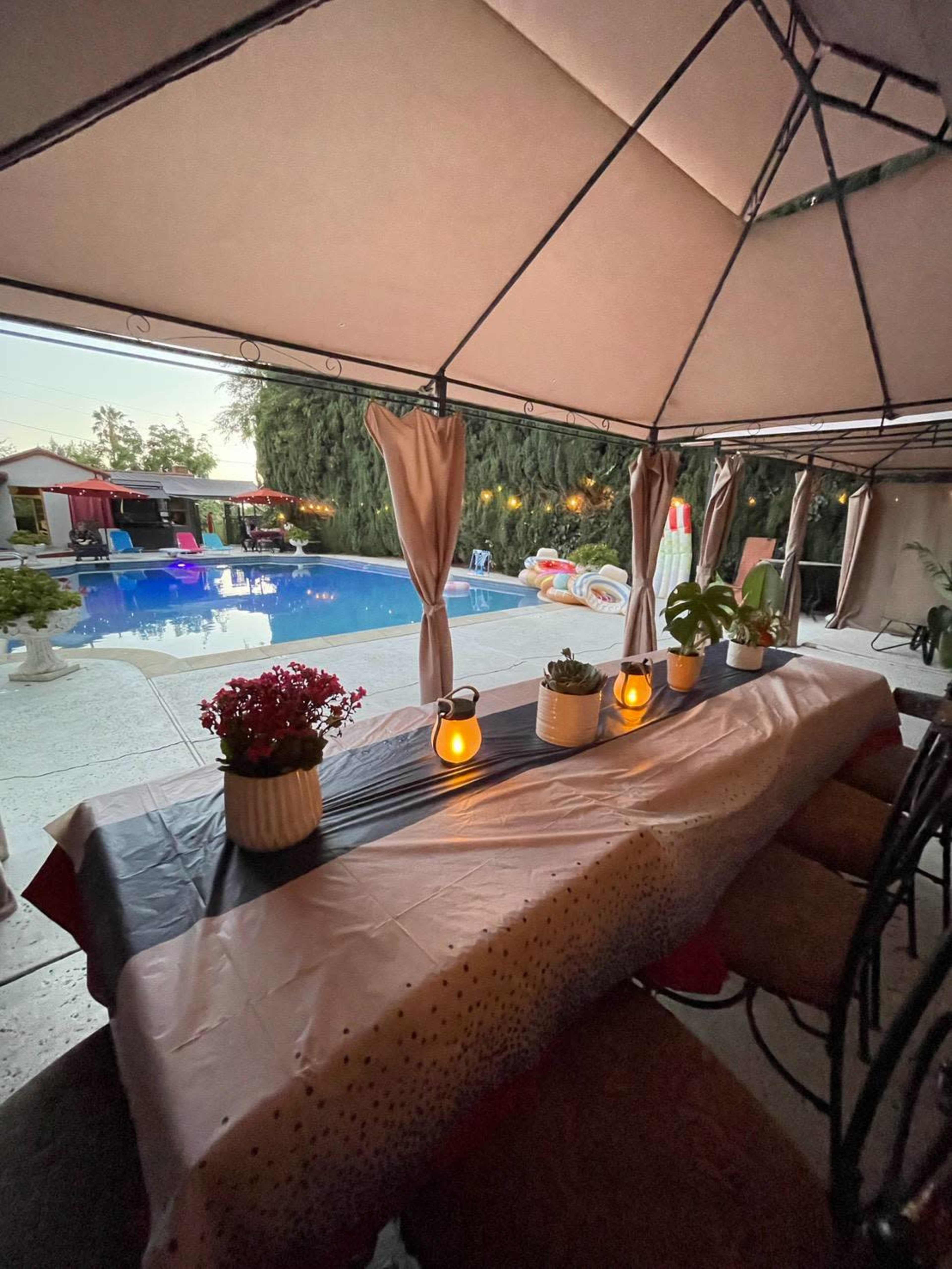 The image shows a covered outdoor dining area with a table set up beside a swimming pool, surrounded by plants and lanterns.