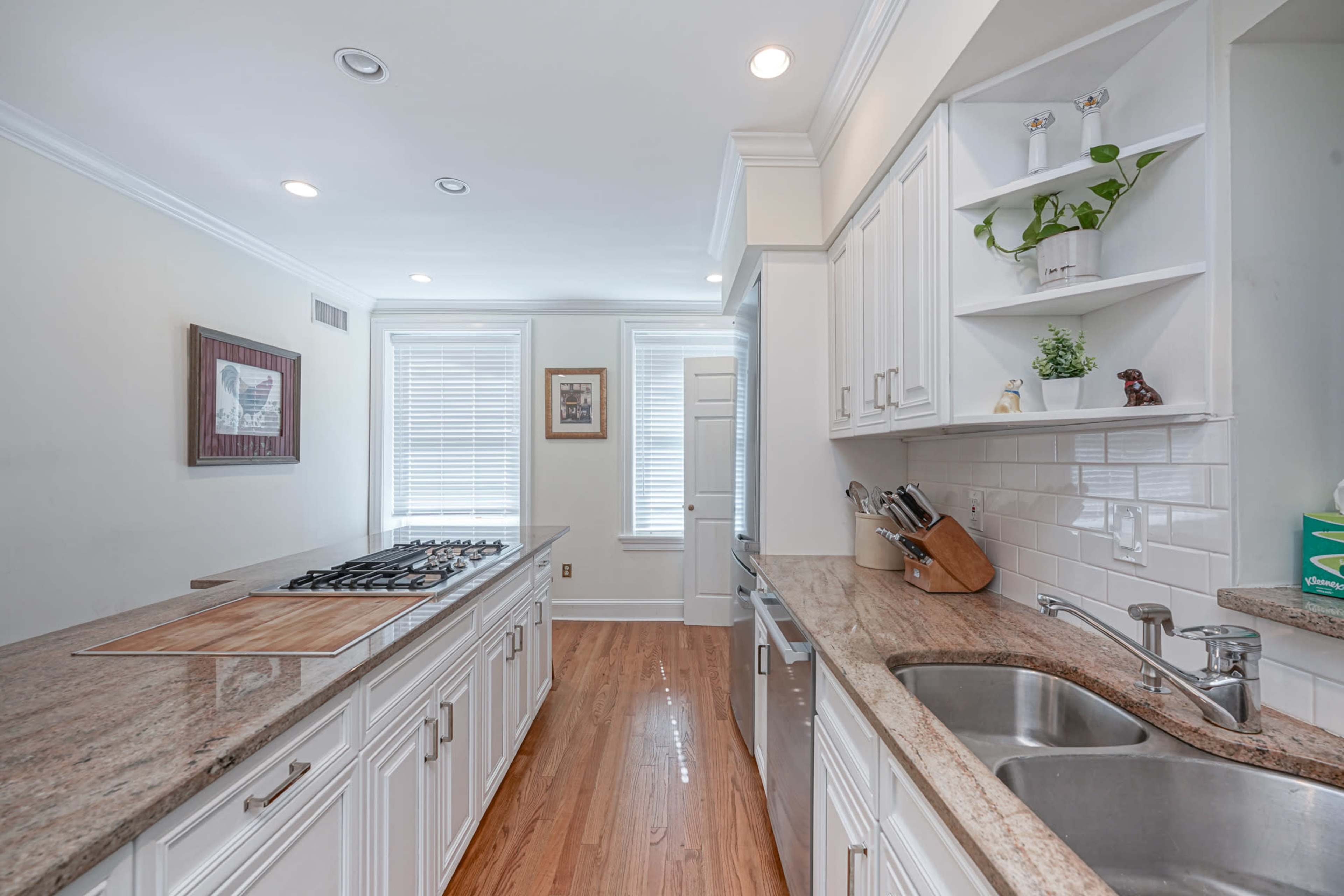 The image shows a modern kitchen with white cabinetry, a granite countertop, a gas cooktop, and a double sink, illuminated by natural light from windows.
