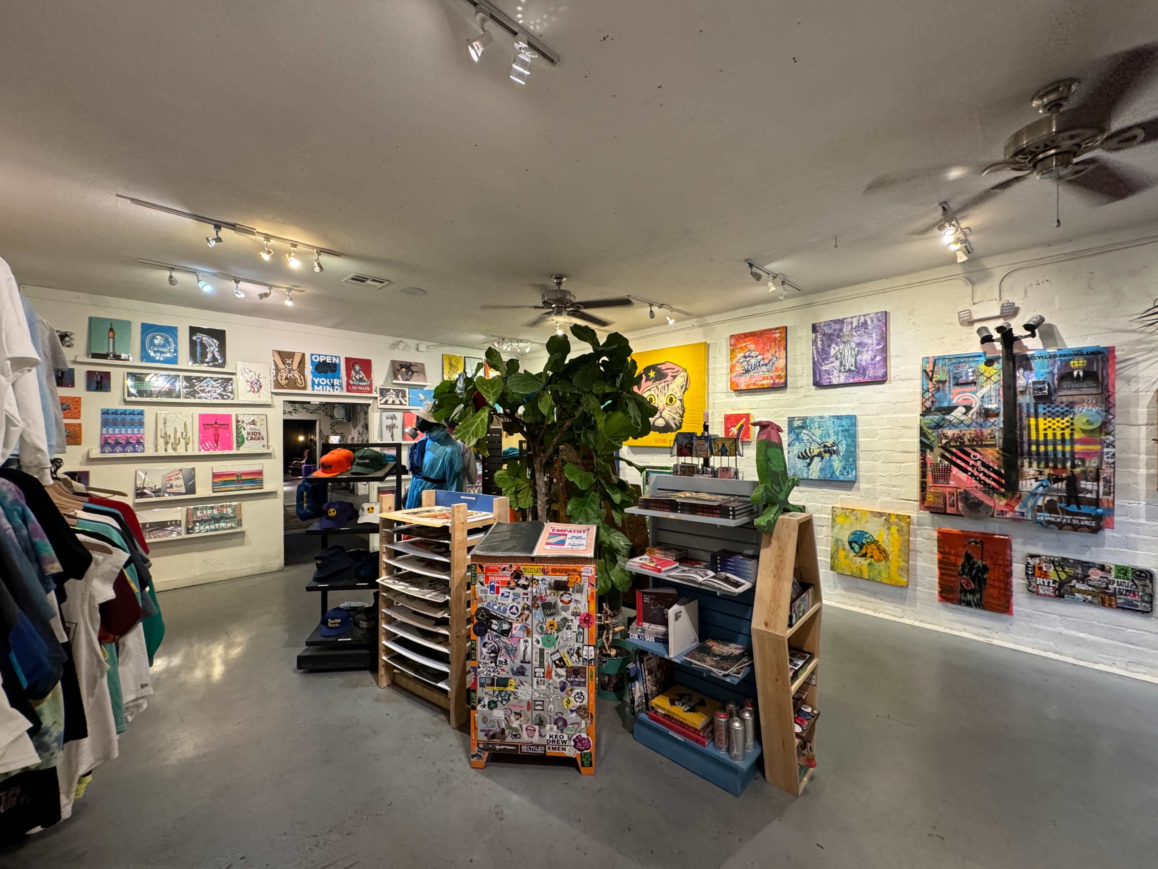 The image shows an art studio with colorful artwork displayed on the walls, a central display rack, and various items for sale, including clothing and art supplies.