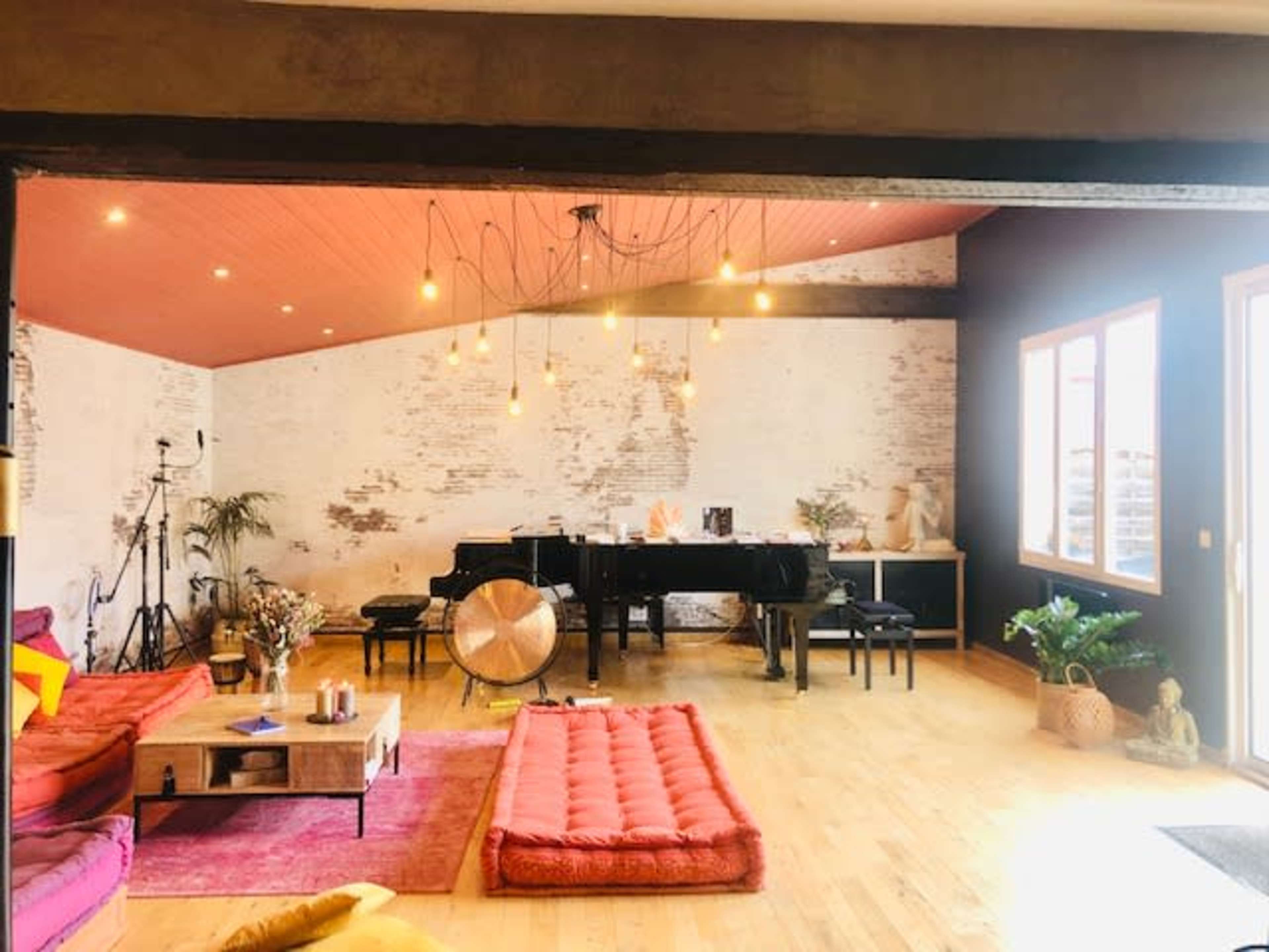 The image shows a spacious room with a grand piano, multiple seating areas, and warm lighting fixtures, complemented by wooden floors and decorative plants.