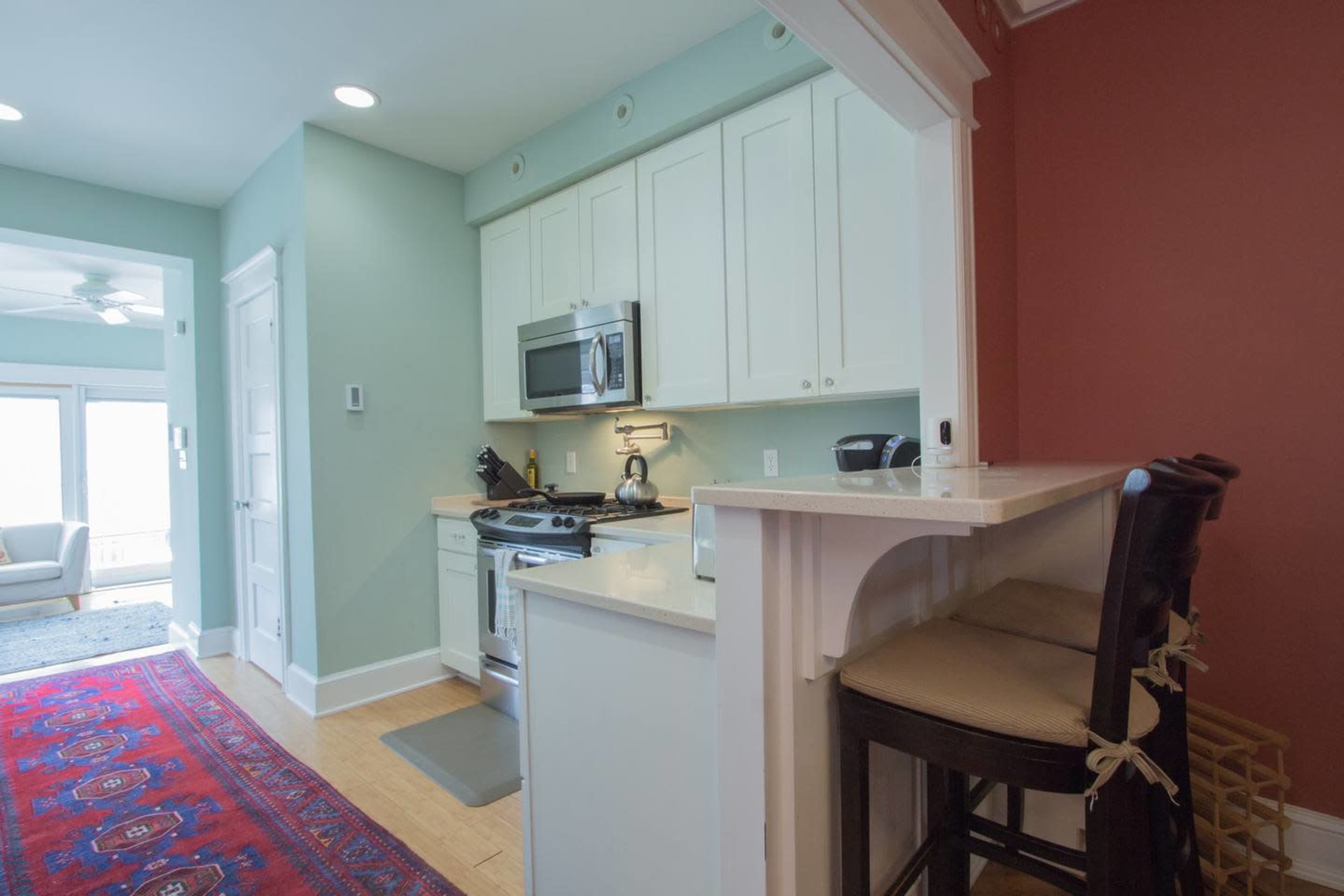 A small kitchen area with white cabinets, a stainless steel microwave, and a bar with two dark stools.