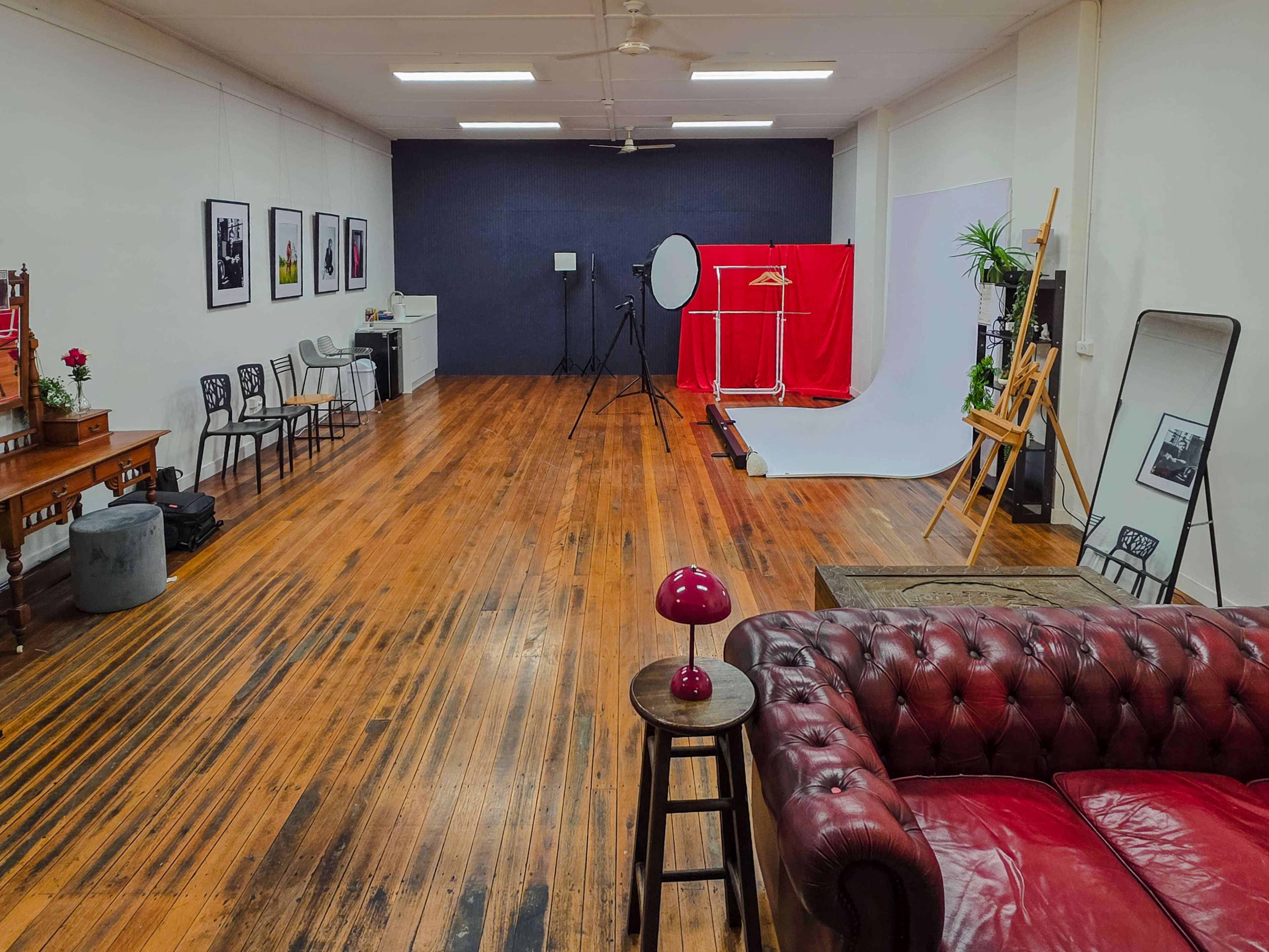 The image shows a spacious studio with polished wooden floors, a red backdrop, photography equipment, and seating arrangements, including a red leather couch and a small table with a lamp.