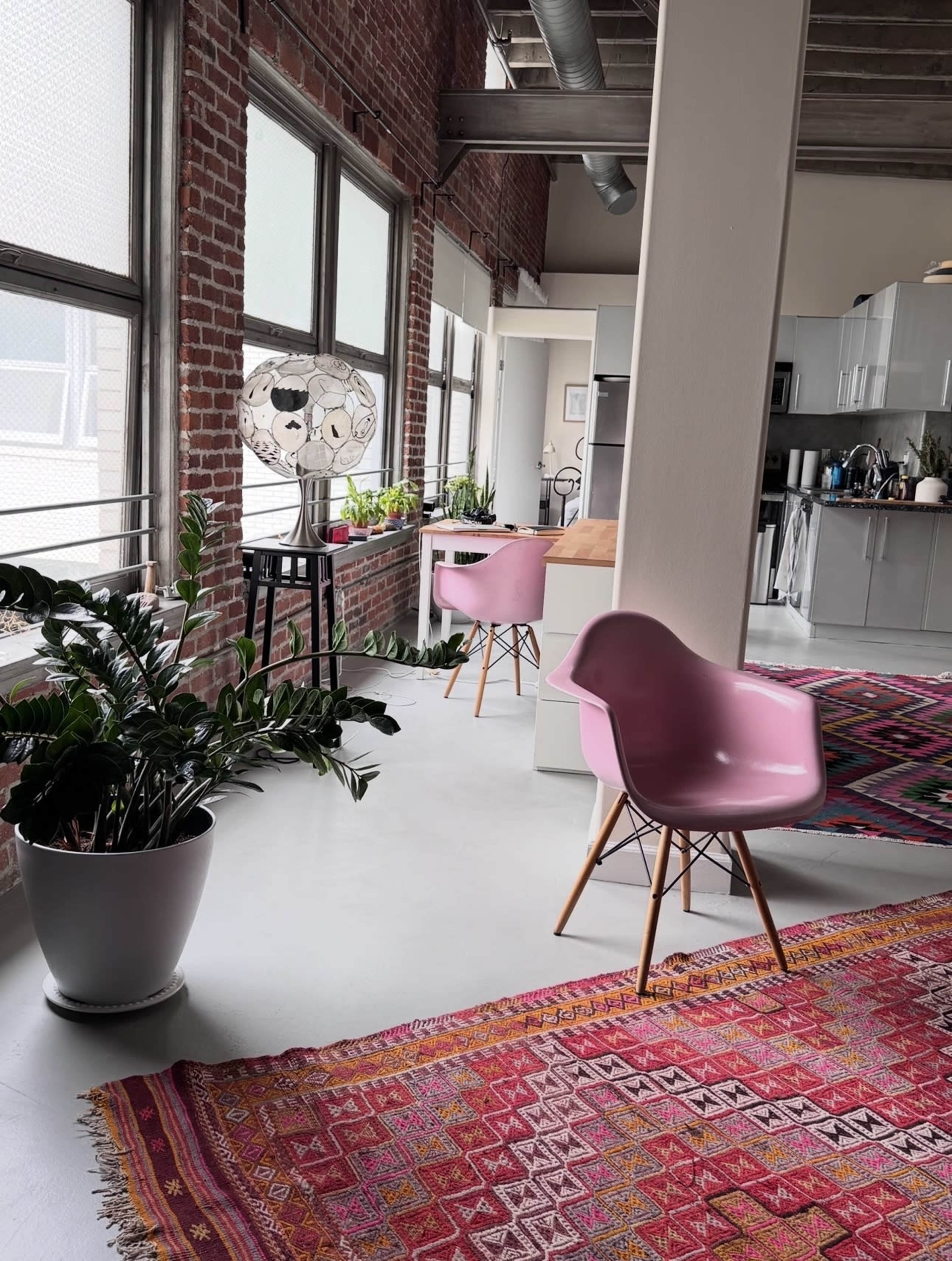 The image shows a modern, open living space with large windows, a pink chair, and a patterned rug against a backdrop of exposed brick walls and a kitchen area.