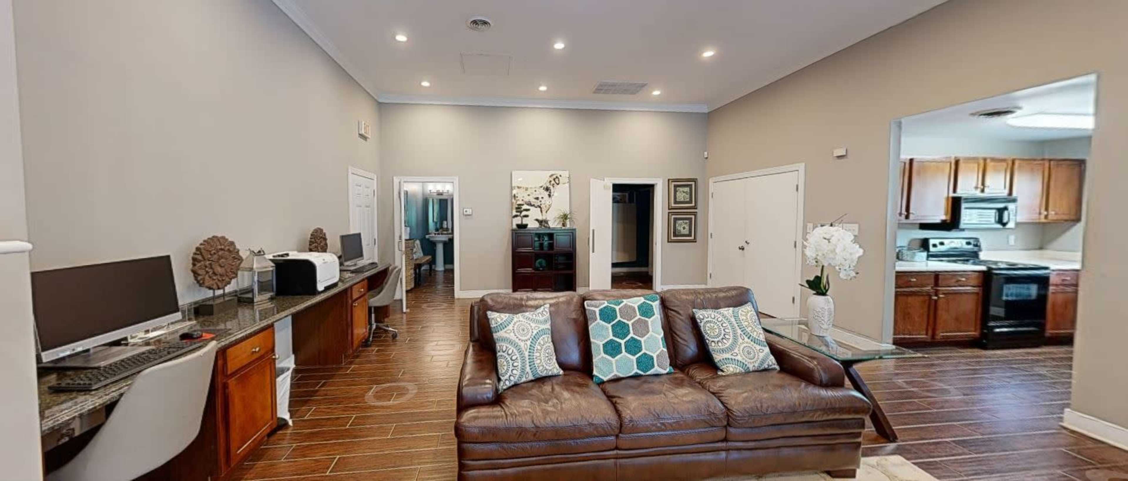 A modern living room with a brown leather sofa, a desk with a computer, and a view of a kitchen area in the background.